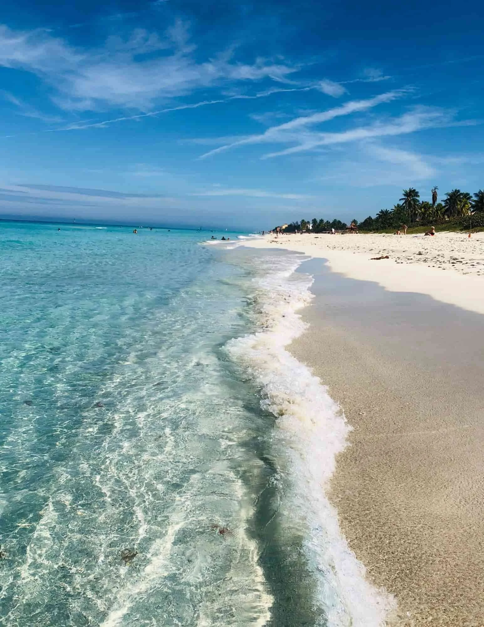 Tropical beach with clear turquoise water, gentle waves, and white sand, with people in the distance and palm trees lining the shore.