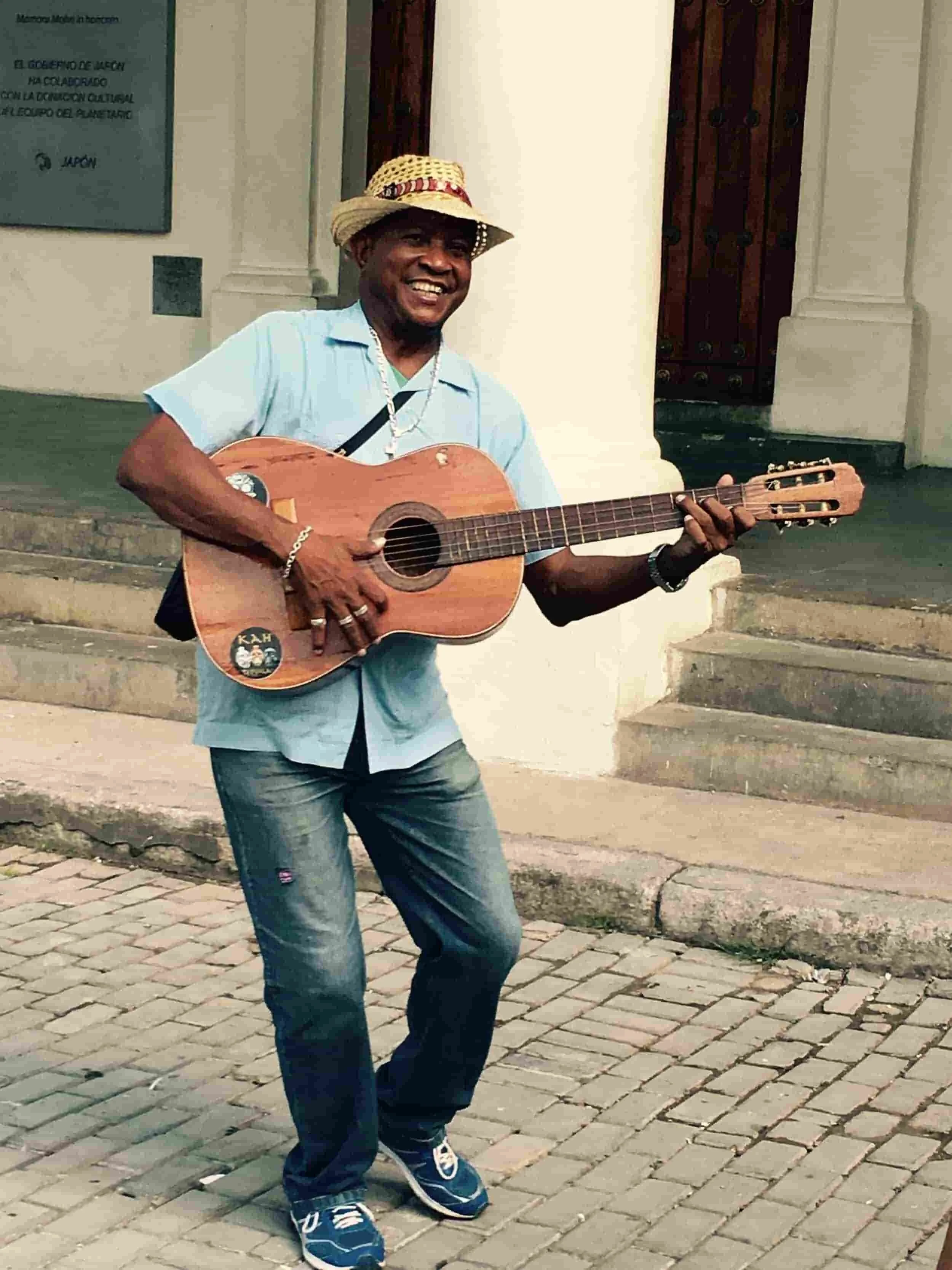 Man in a straw hat smiling while holding a guitar outside a building.