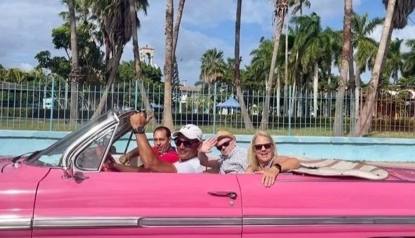 A group of people enjoying a ride in a pink vintage convertible car with palm trees in the background.