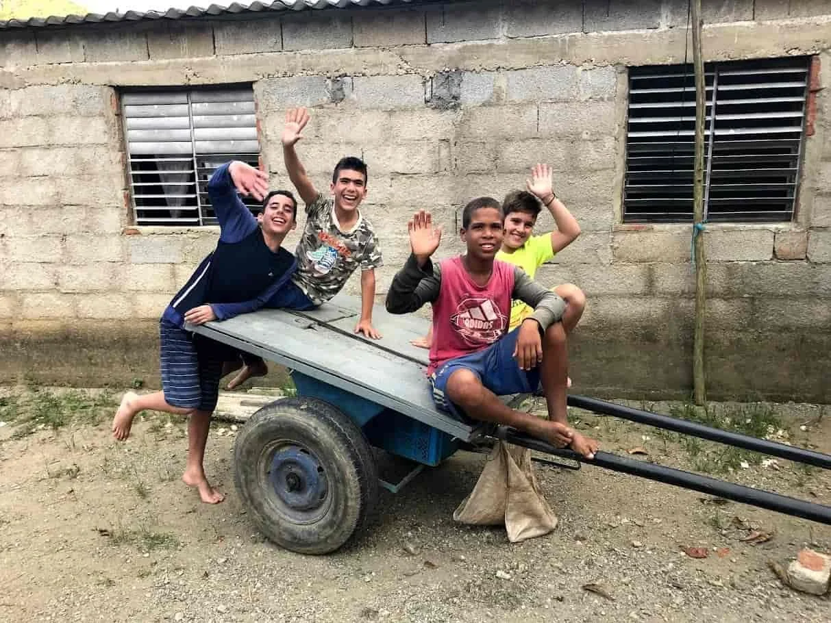 Four boys on cart smiling and waving goodbye outside a gray concrete house with barred windows.