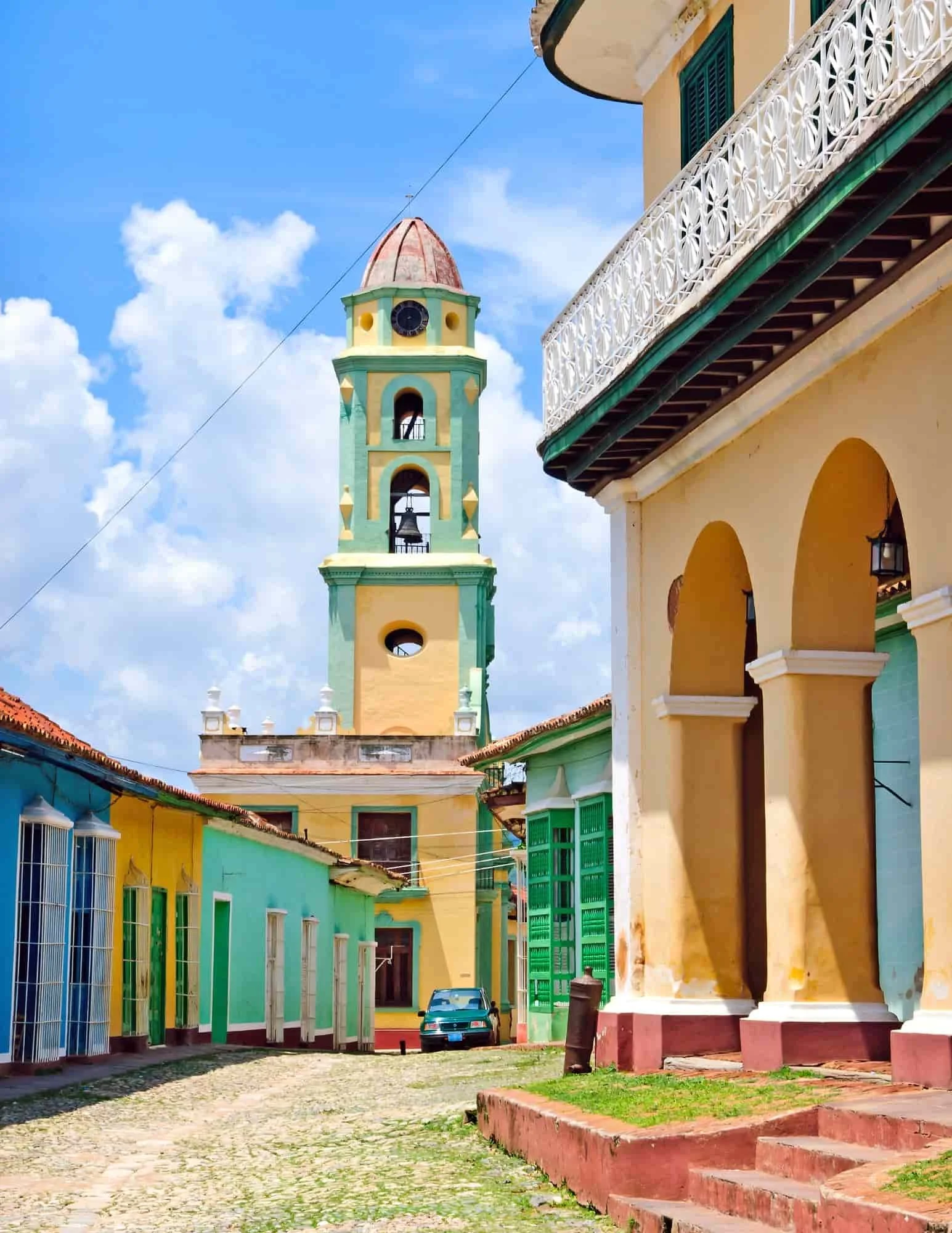 A cobblestone street with colorful colonial buildings, a colorful bell tower, and an old car parked in the distance under a blue sky.