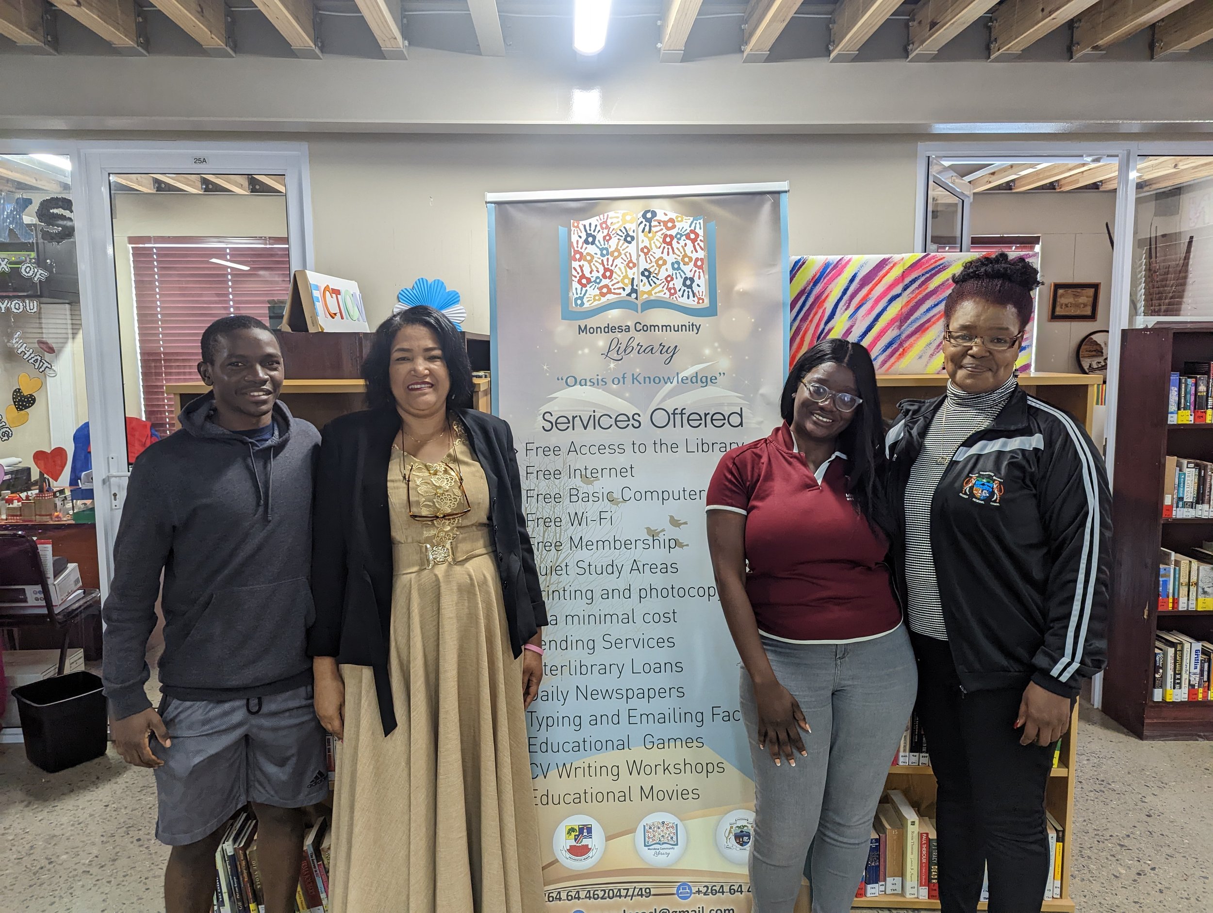 Four people standing in a library next to a sign listing services offered, such as free access and internet.