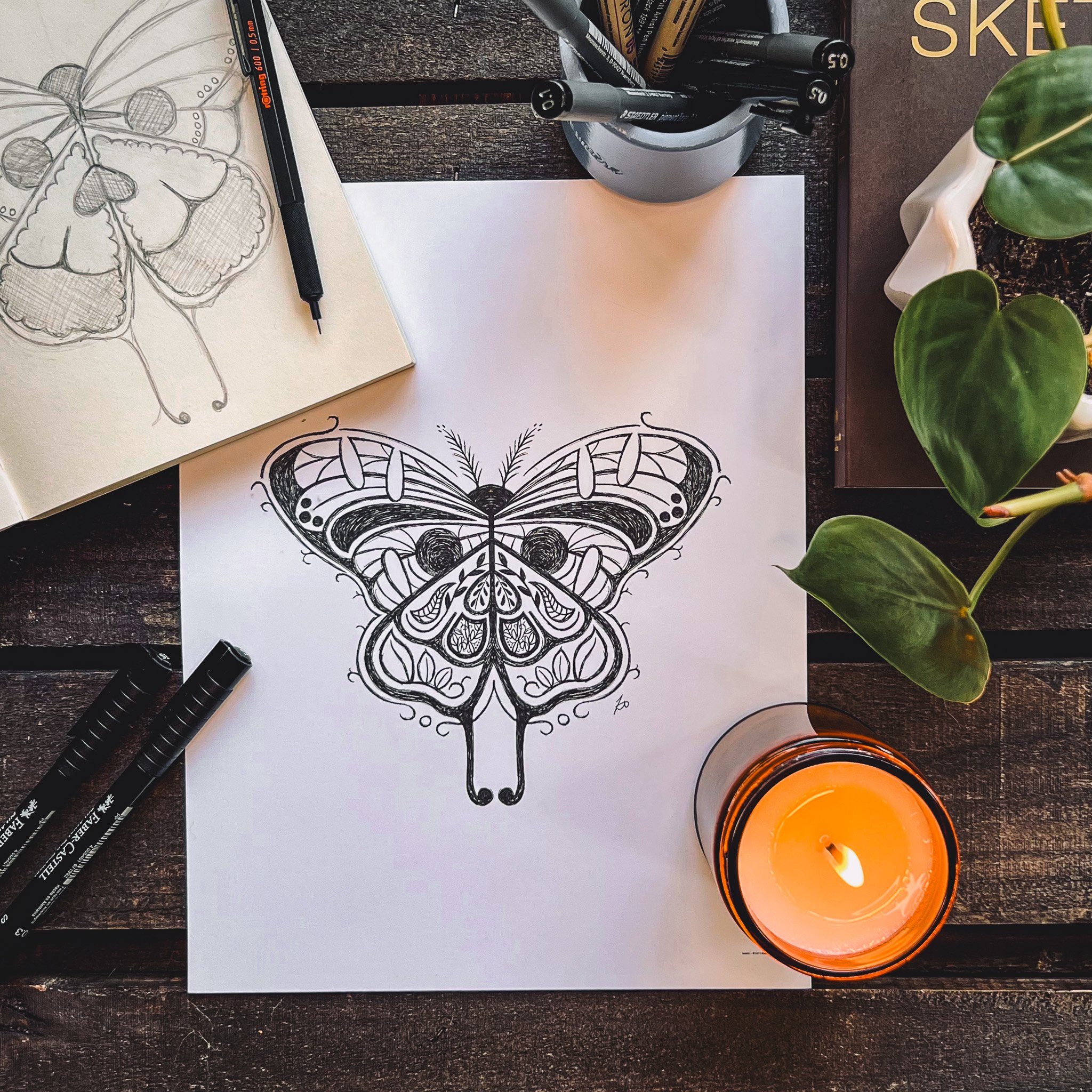 Birdseye view of final print of Larry the moth with ink pen, candle, leaves, and skeleton hand next to it on dark wooden background.
