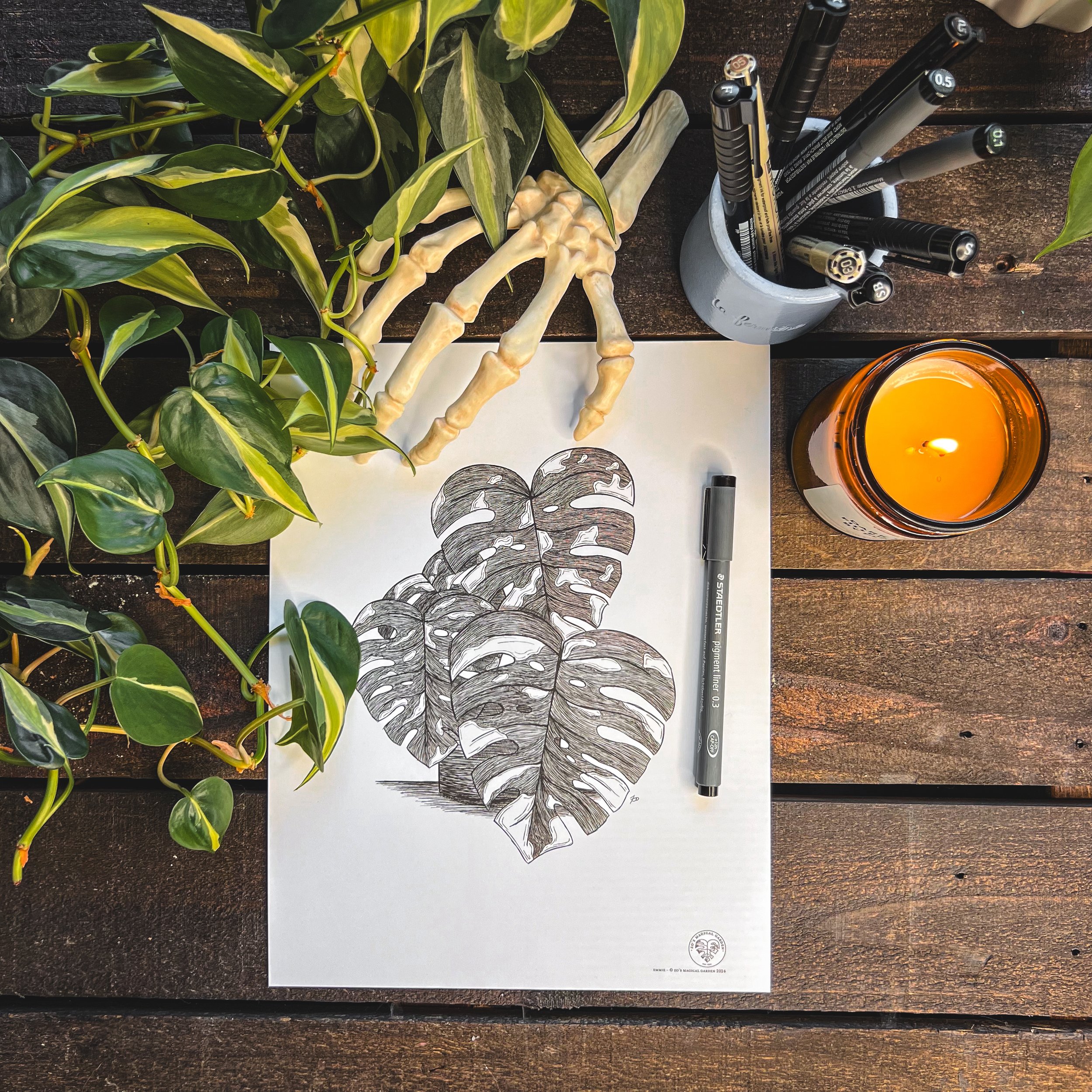 Birdseye view of final print of Emmie the Thai Monstera with ink pen, candle, leaves, and skeleton hand next to it on dark wooden background.