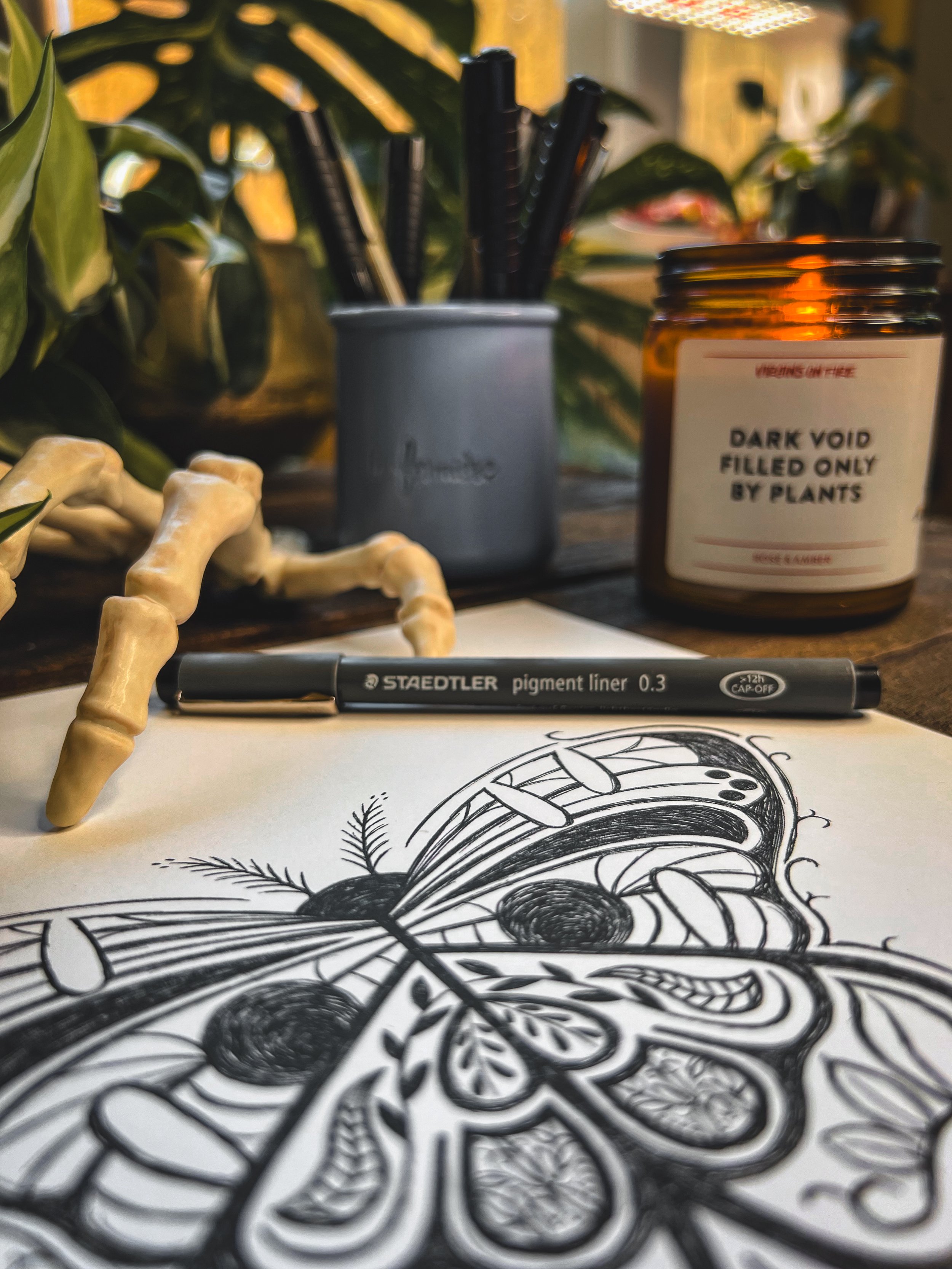 Final print of Larry the moth with ink pen, candle, leaves, and skeleton hand next to it on dark wooden background.