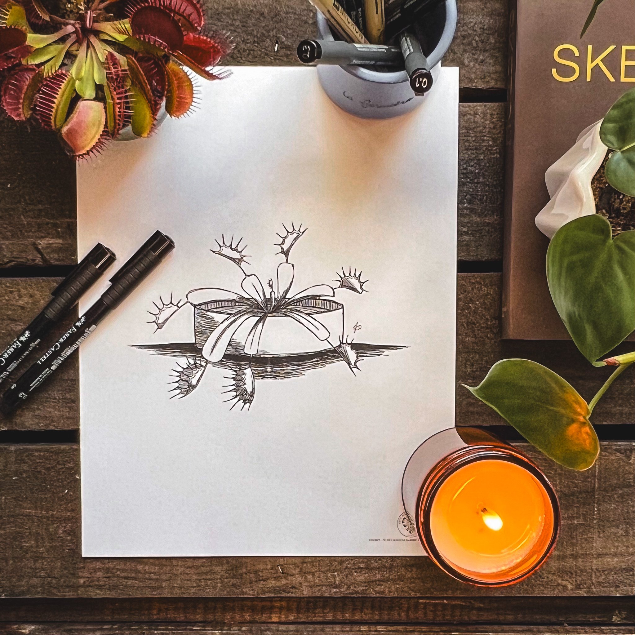 birdseye view of final print of Chompy the venus fly trap. Ink pens, candle, fly trap and sketchbook on a dark wooden background surrounding the print.