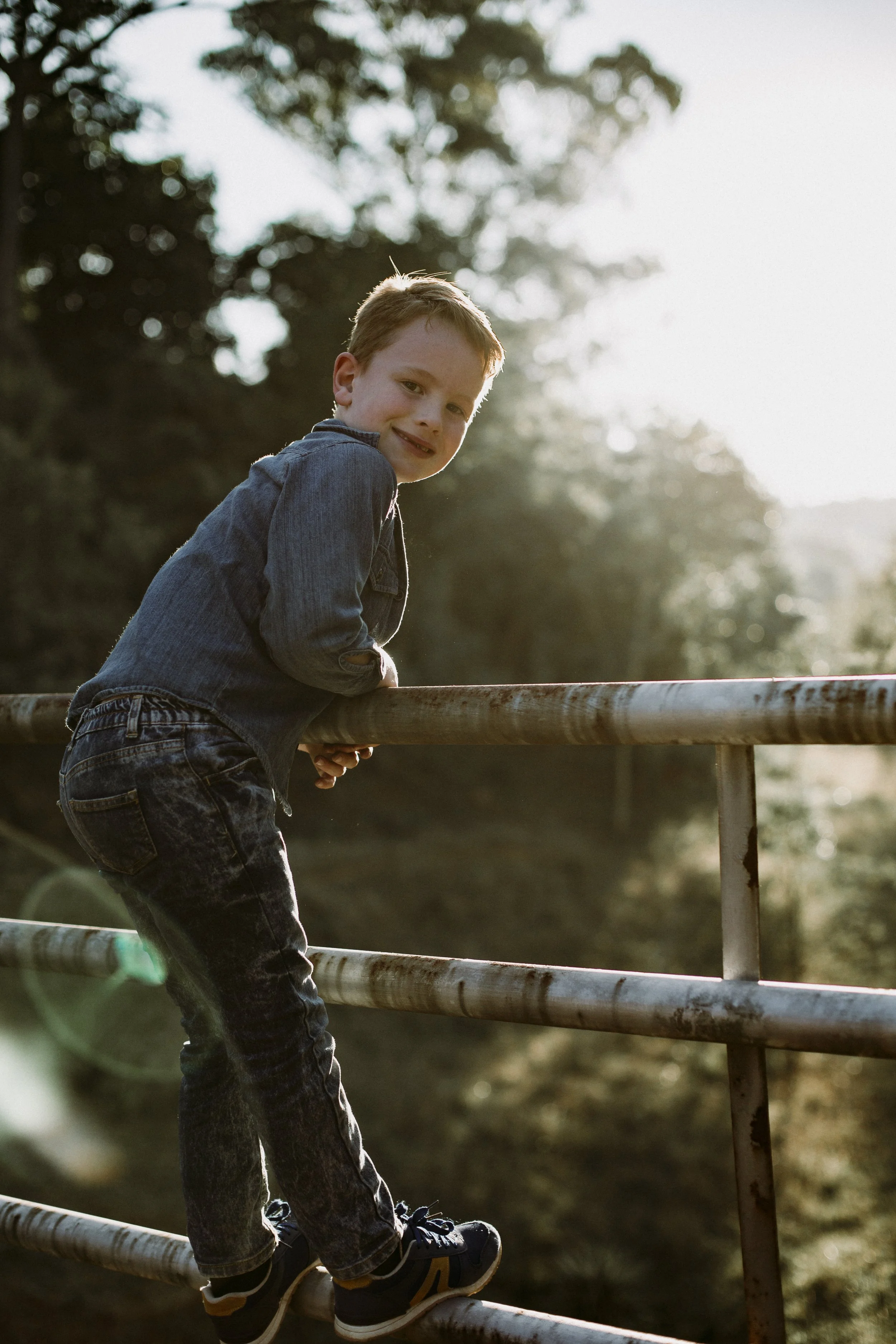 Young boy in denim jacket and jeans balancing on a rusty metal railing outdoors during sunset, smiling at the camera with trees and sky in the background.