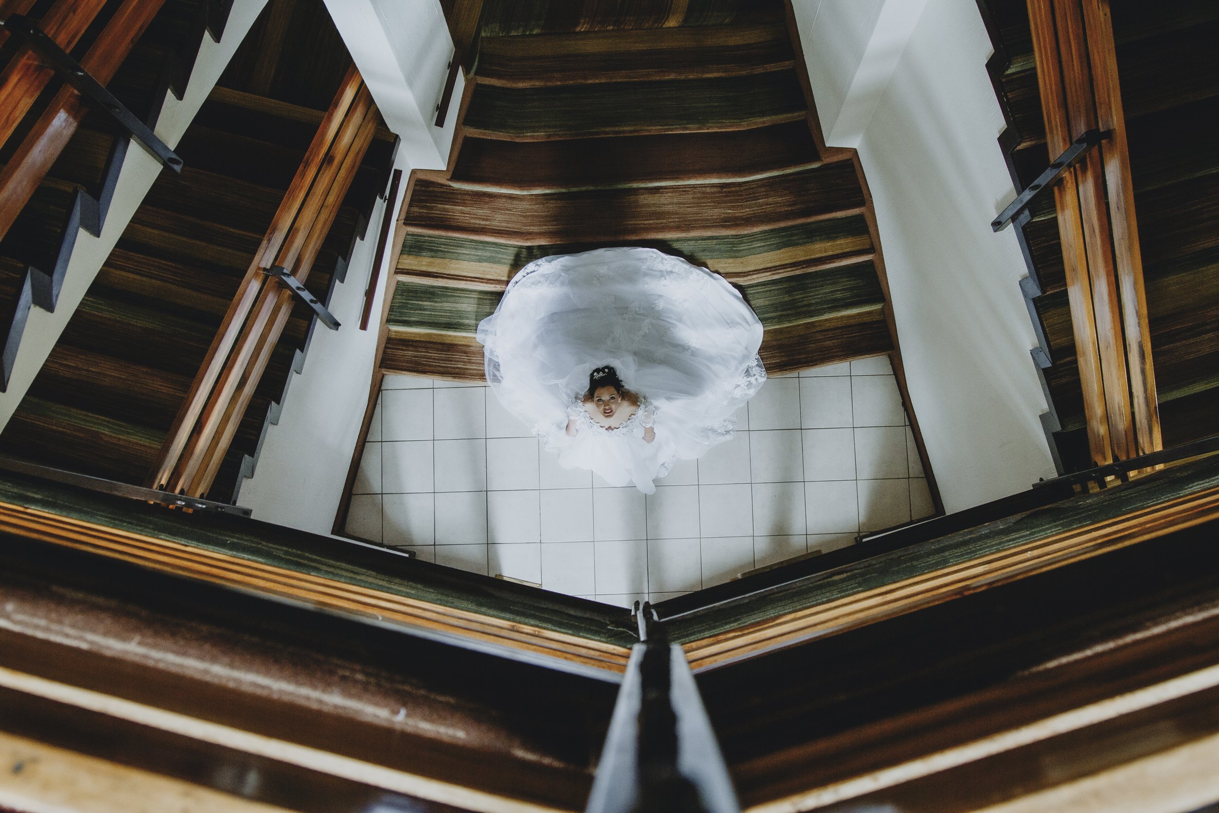 Top-down view of a young girl in a white wedding dress looking up from the bottom of a staircase with wooden rails and tiled floor.
