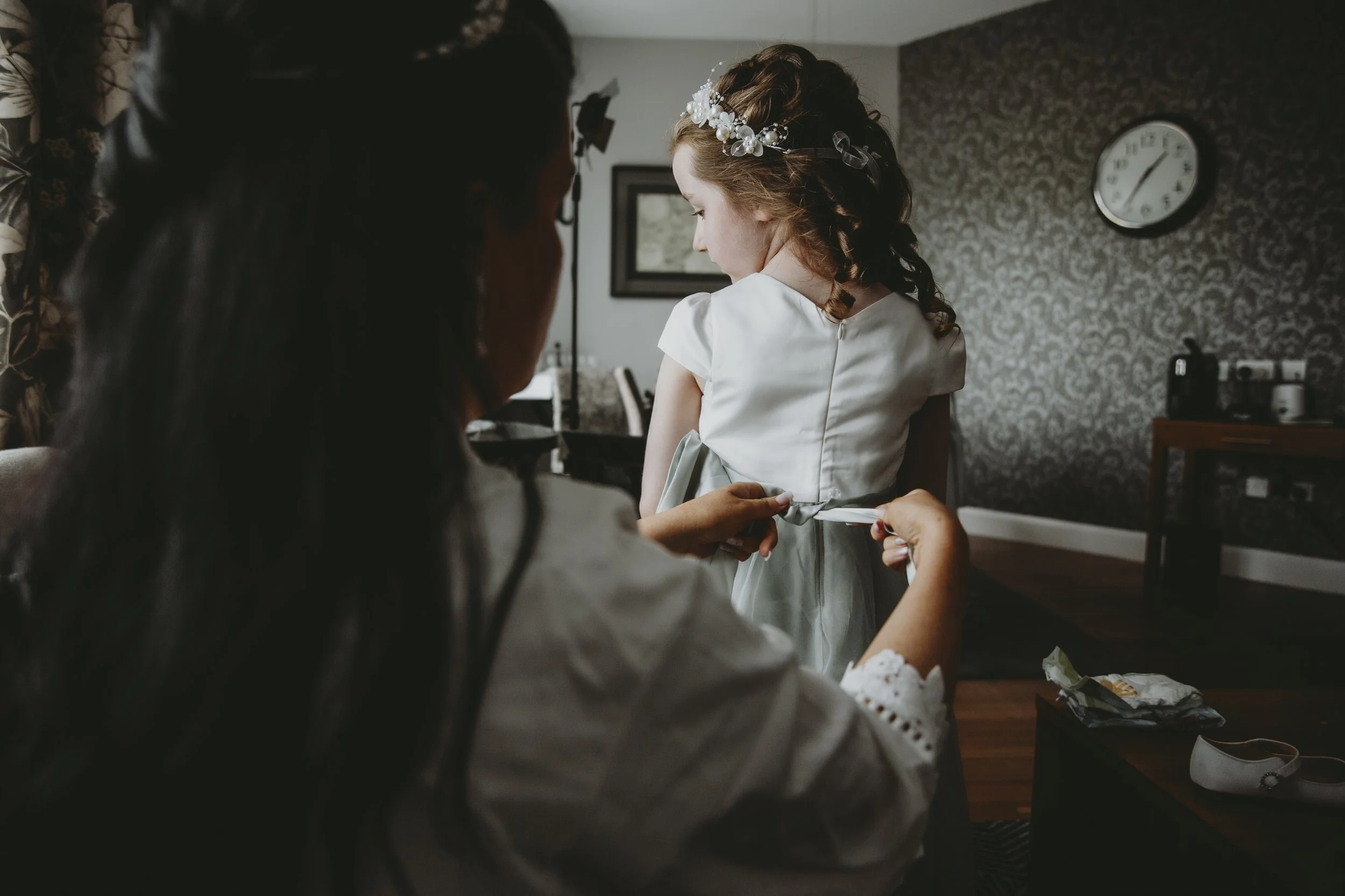 A woman helps a young girl put on a ribbon at the back of her dress in a room with patterned wallpaper and a clock on the wall.