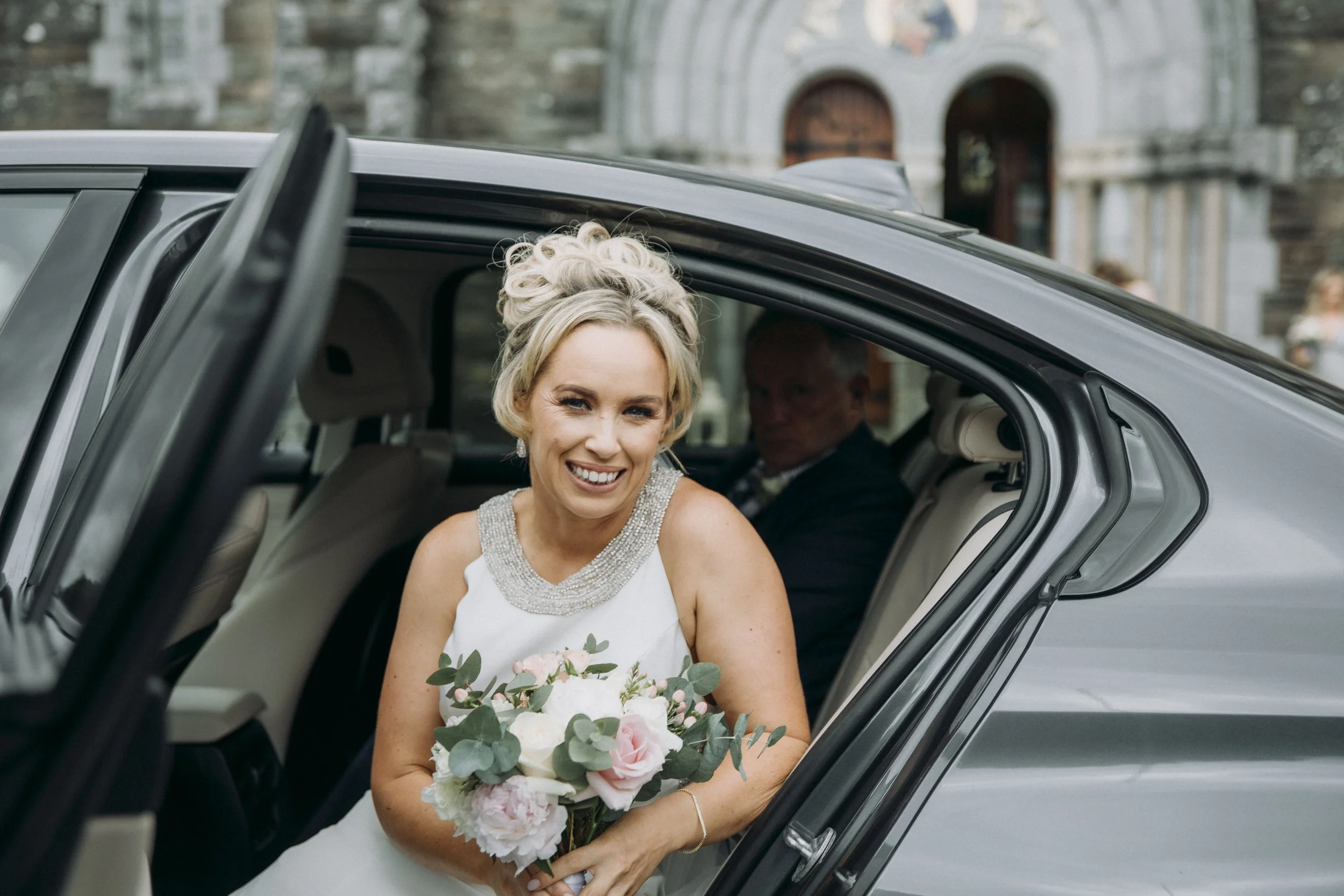 A smiling bride holding a bouquet of flowers sitting in the back seat of a car, with a man in a suit in the background and a stone building in the distance. timoleague abbey
