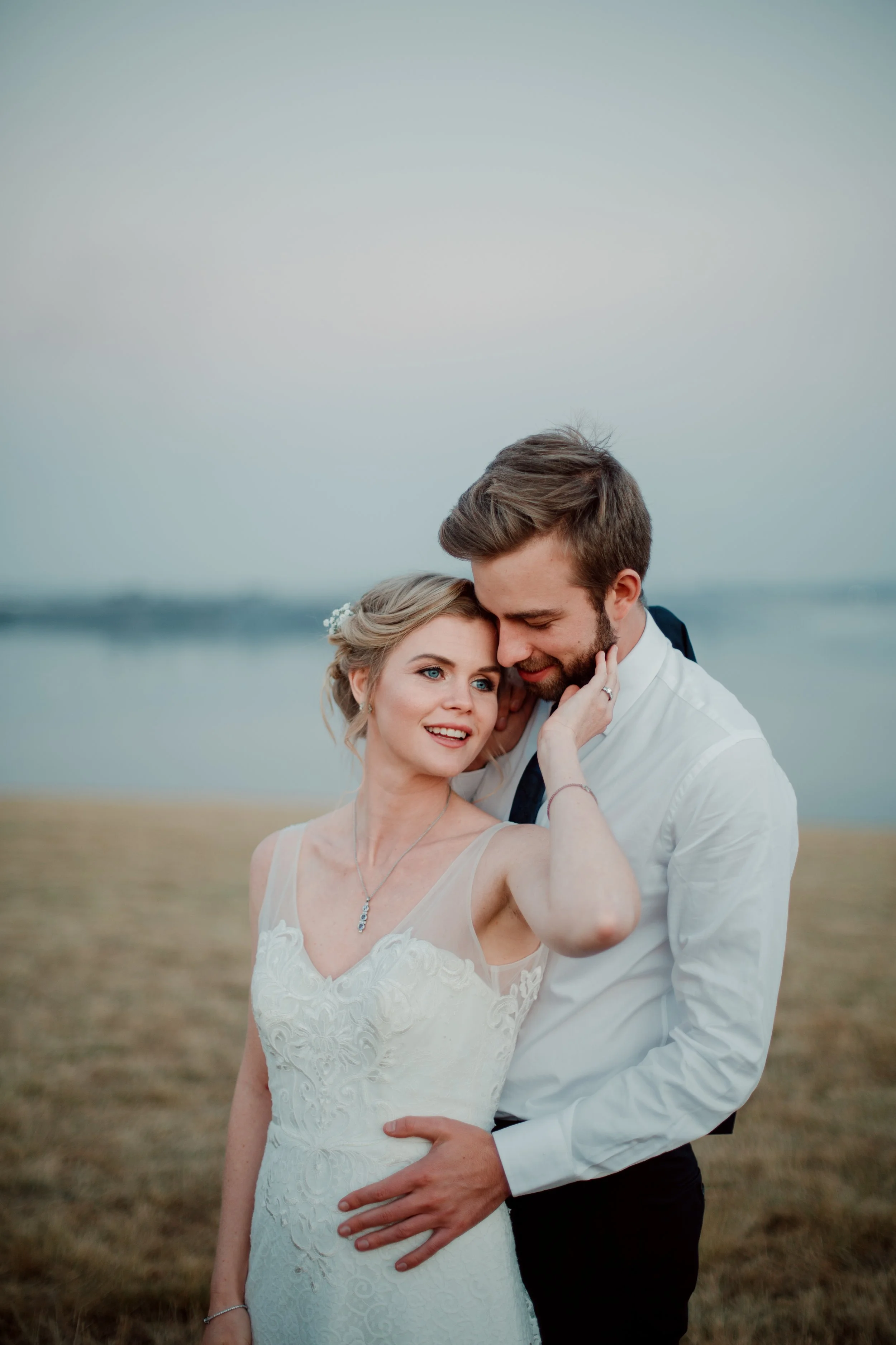 A newlywed couple sharing an affectionate moment outdoors near a body of water, dressed in wedding attire. The bride is in a white lace gown, and the groom is in a white shirt with dark trousers.
