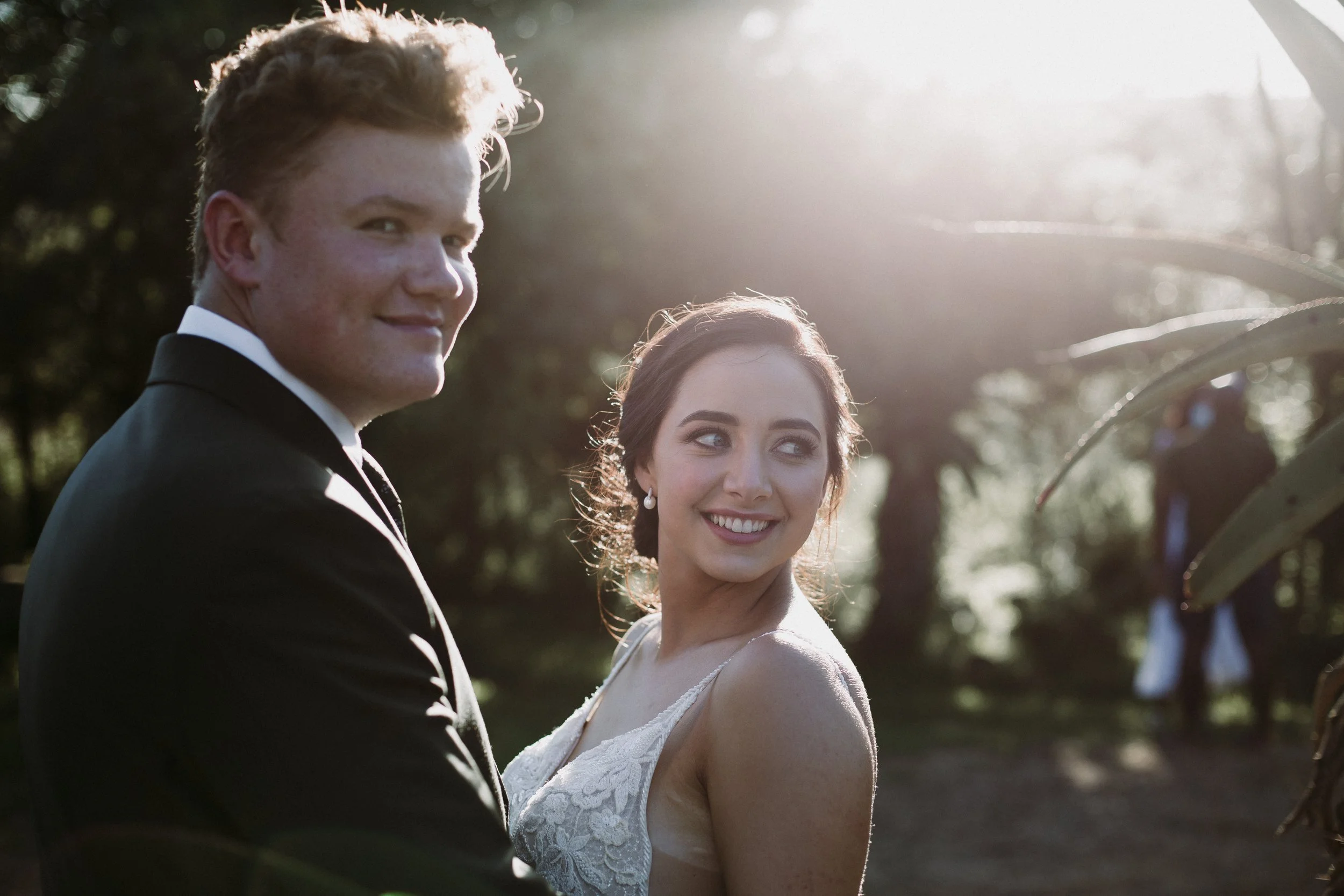 A bride and groom stand together outdoors at sunset, smiling and looking at each other, with trees in the background.