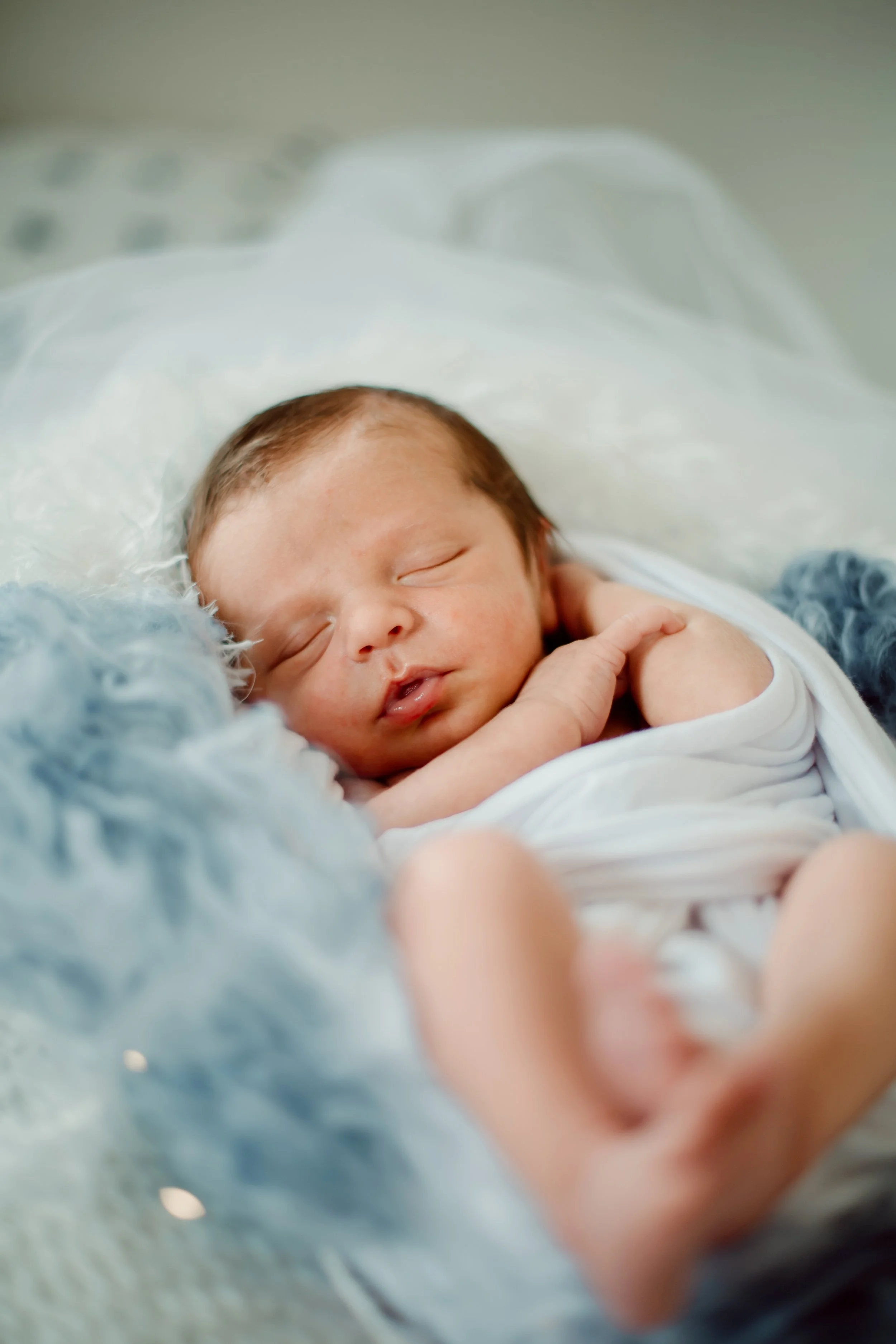A sleeping newborn baby with light skin and brown hair, wrapped in a white blanket, nestled on a soft, fuzzy blue blanket.