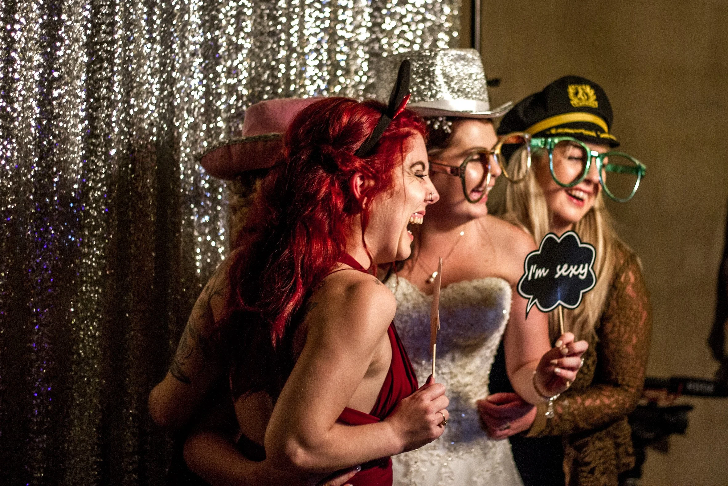 Three women smiling and laughing at a party, wearing fun hats and oversized glasses, holding signs that say 'I'm sexy'. One woman has red hair, the other two have blonde hair, and they are standing in front of a sparkly silver backdrop.