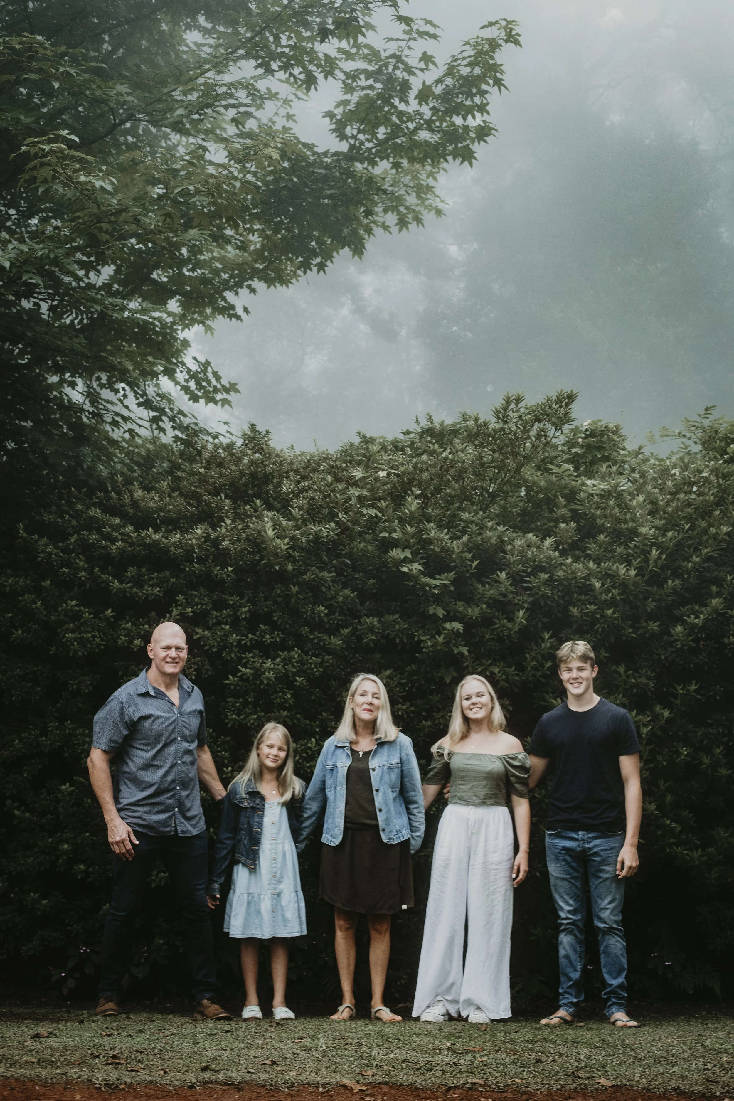 Family portrait of five people standing outdoors on grass, with tall lush greenery and foggy trees in the background.