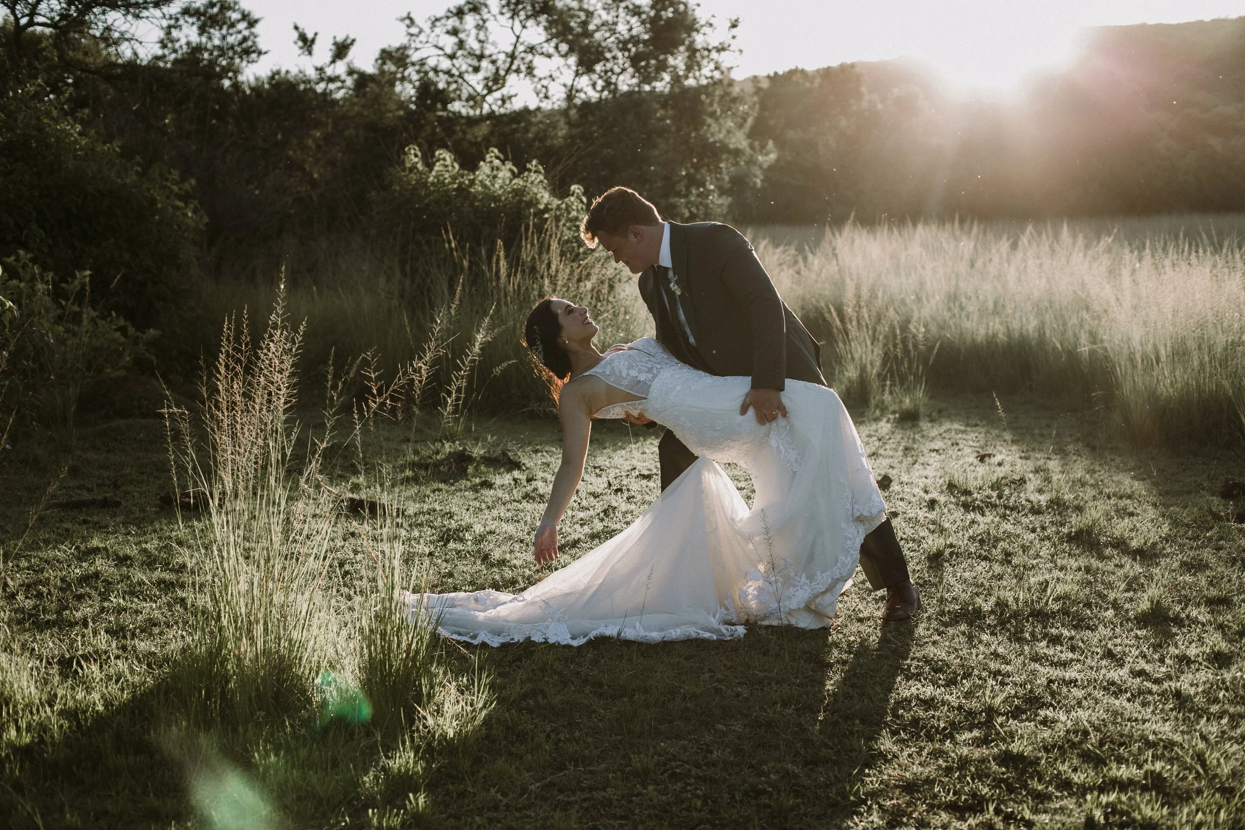 A wedding couple dancing outdoors at sunset, with the man in a black suit holding the woman in a white wedding dress, surrounded by tall grass and trees.