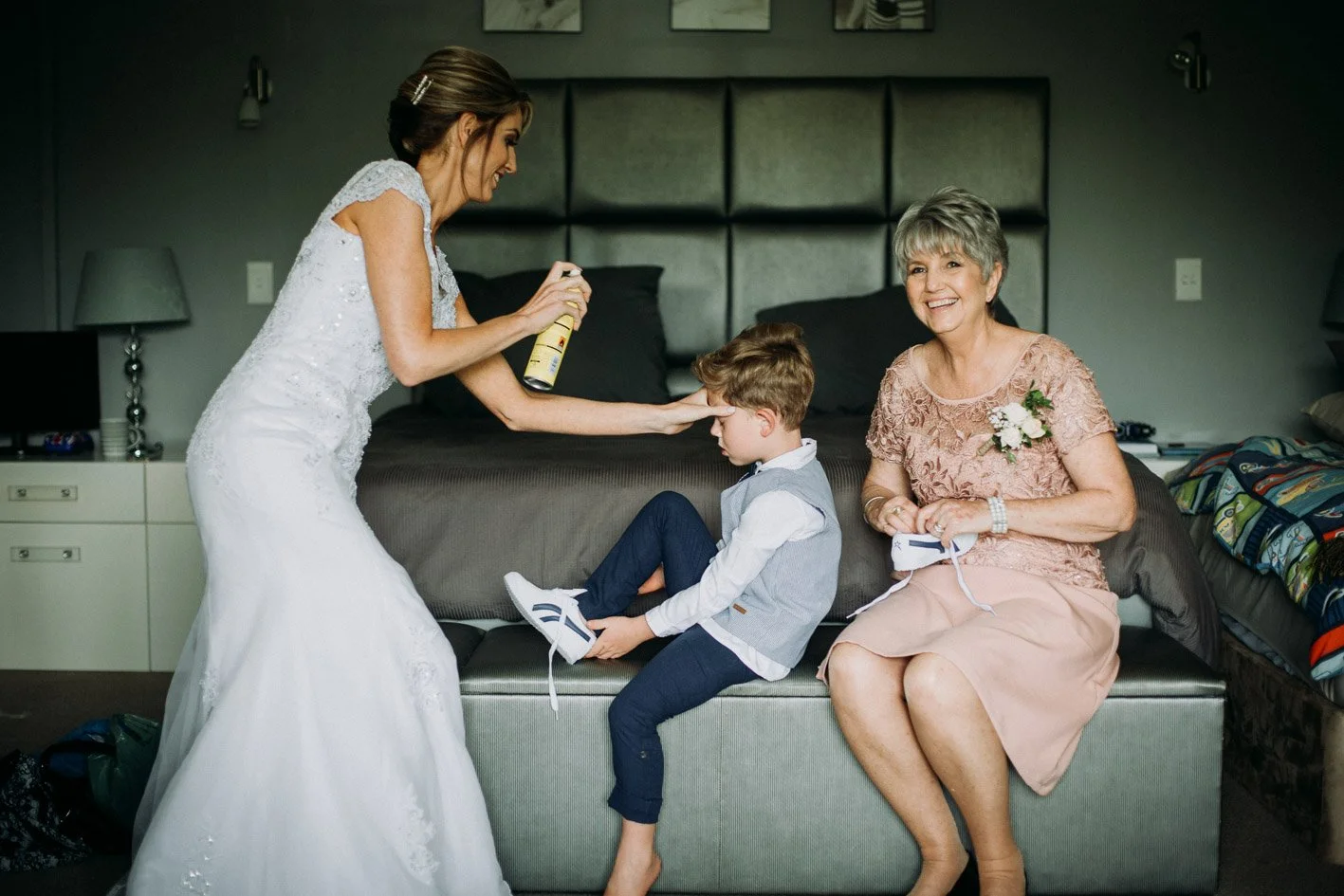 A bride spraying perfume on a young boy's nose, with an elderly woman smiling on a bed in a hotel room.