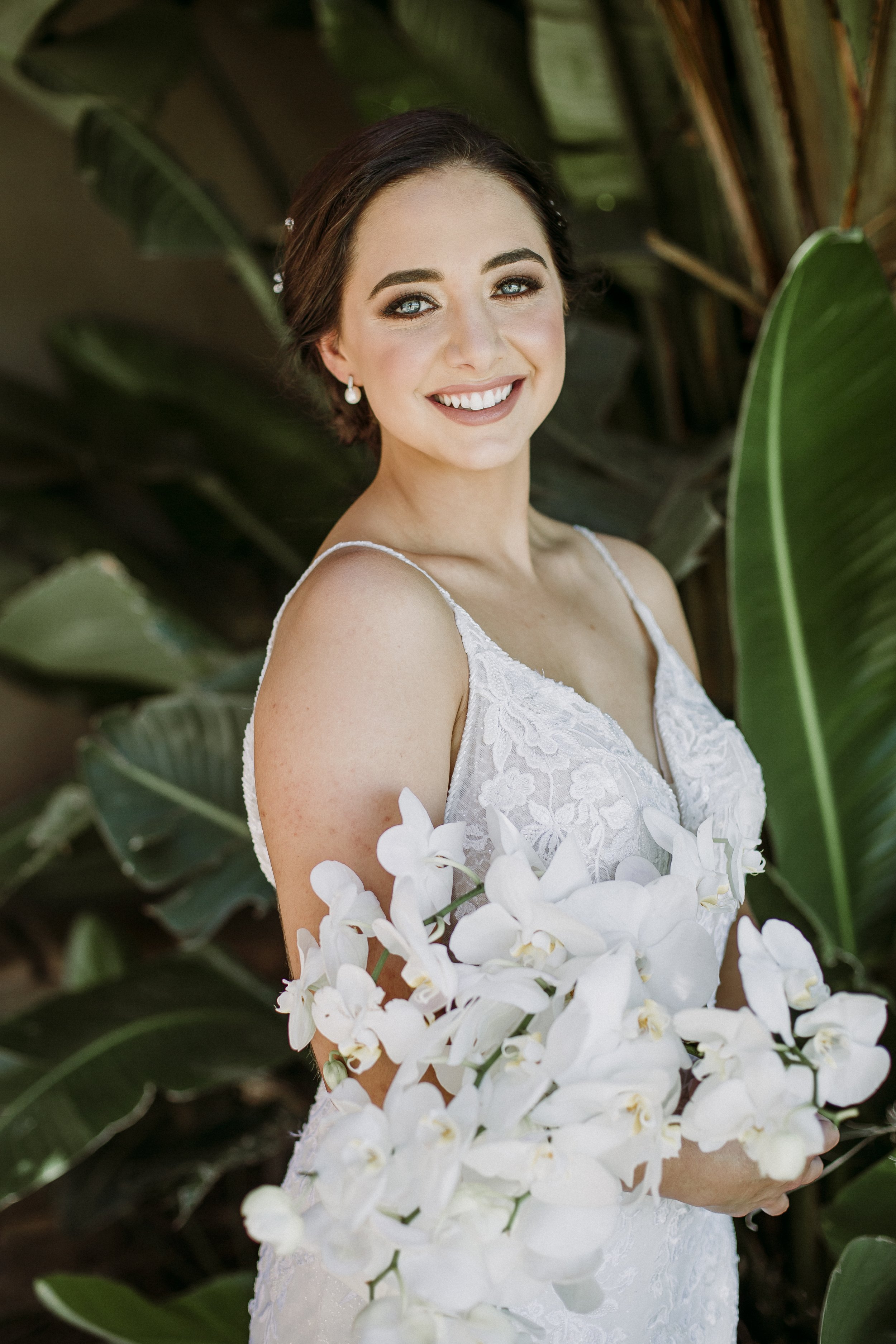 A smiling woman in a white lace wedding dress holding a bouquet of white orchids, standing among large green tropical leaves.