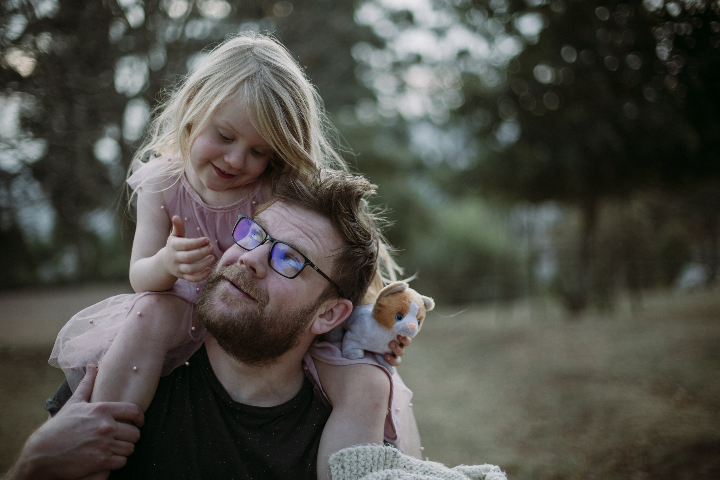 A young girl and a man with glasses are outdoors, with the girl riding on the man's shoulders and touching his face, both smiling; the girl has blonde hair and is wearing a pink dress, and the man has brown hair, a beard, and is wearing a black shirt
