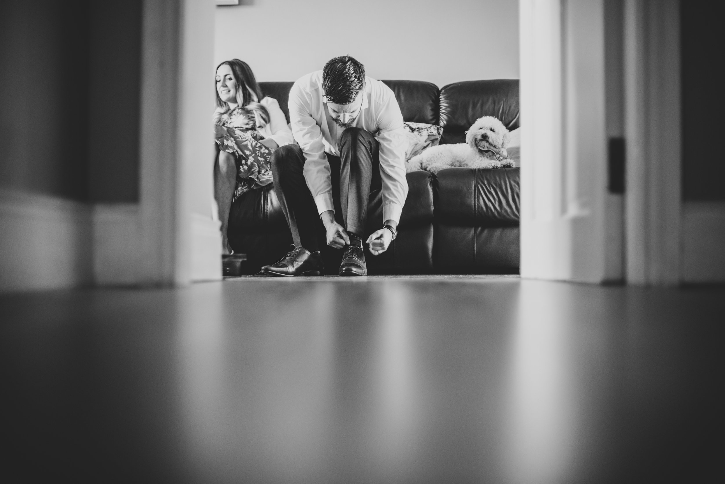 A man is tying his shoes while a woman and a dog sit on a couch behind him. The photo is taken from a low angle, through an open door.