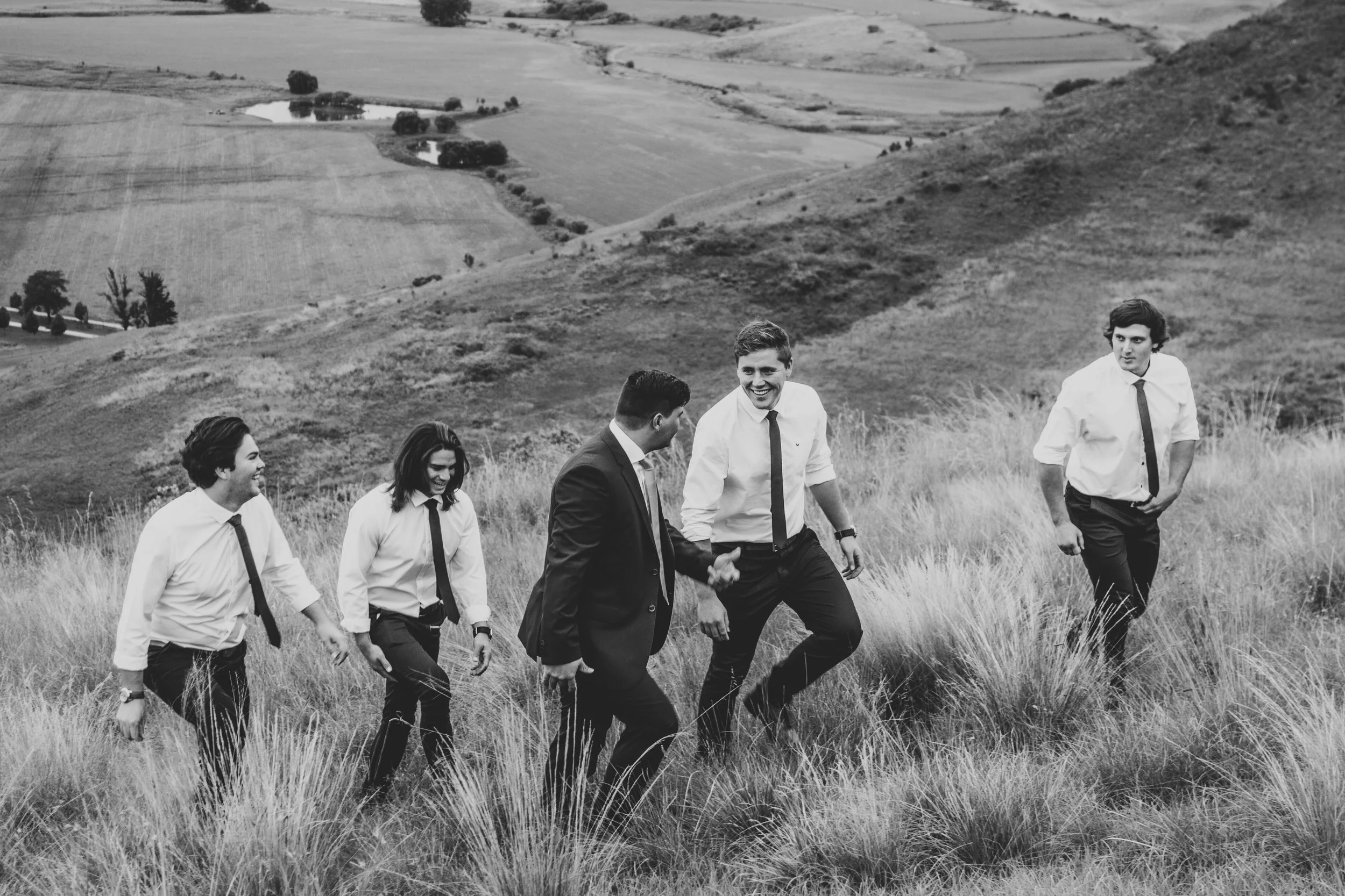 Black and white photo of five men walking on a grassy hillside, dressed in formal shirts and ties, with rolling hills and fields in the background.