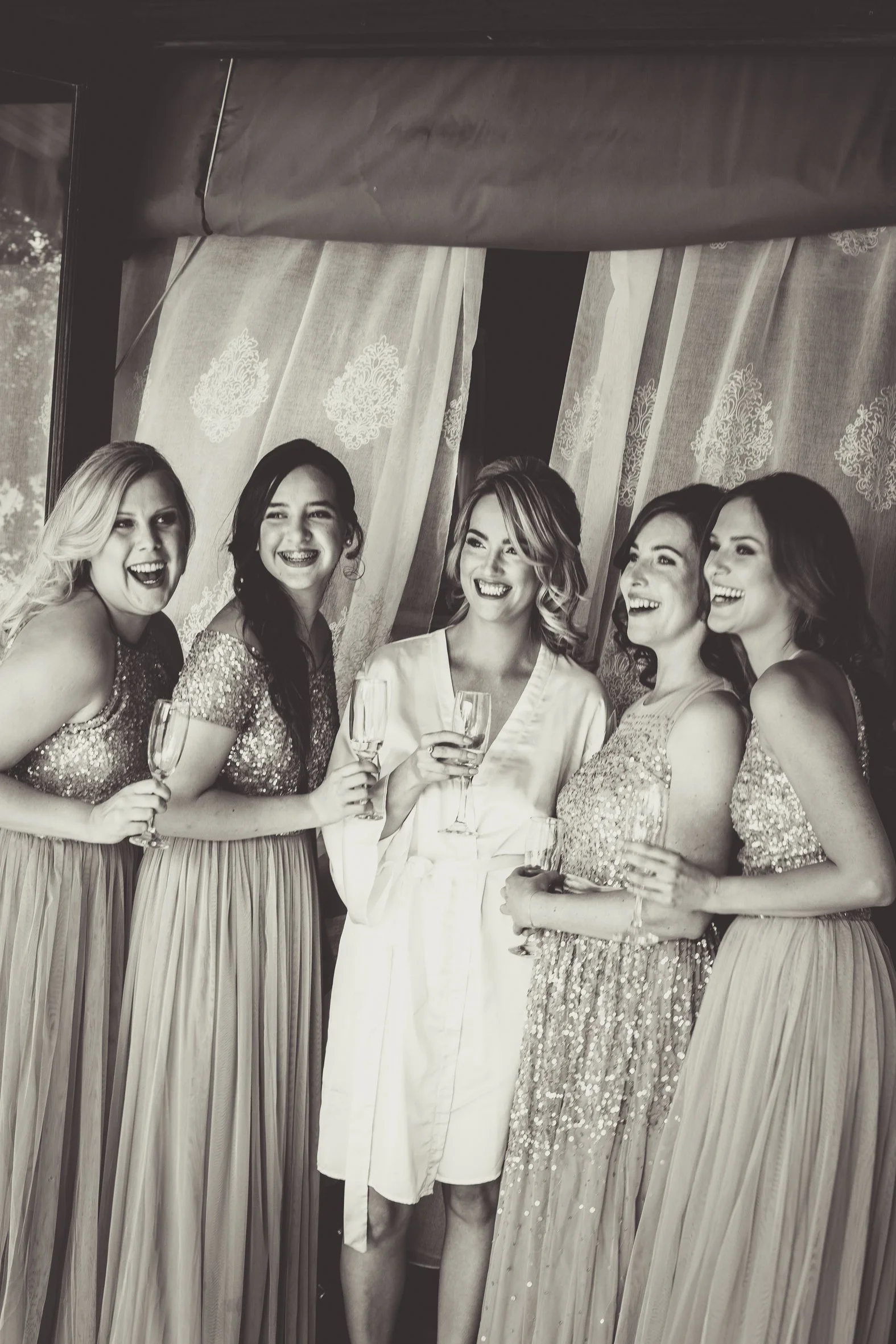 A group of six women, five wearing sparkly dresses with sequins, holding glasses of champagne, smiling and laughing together at a celebration.