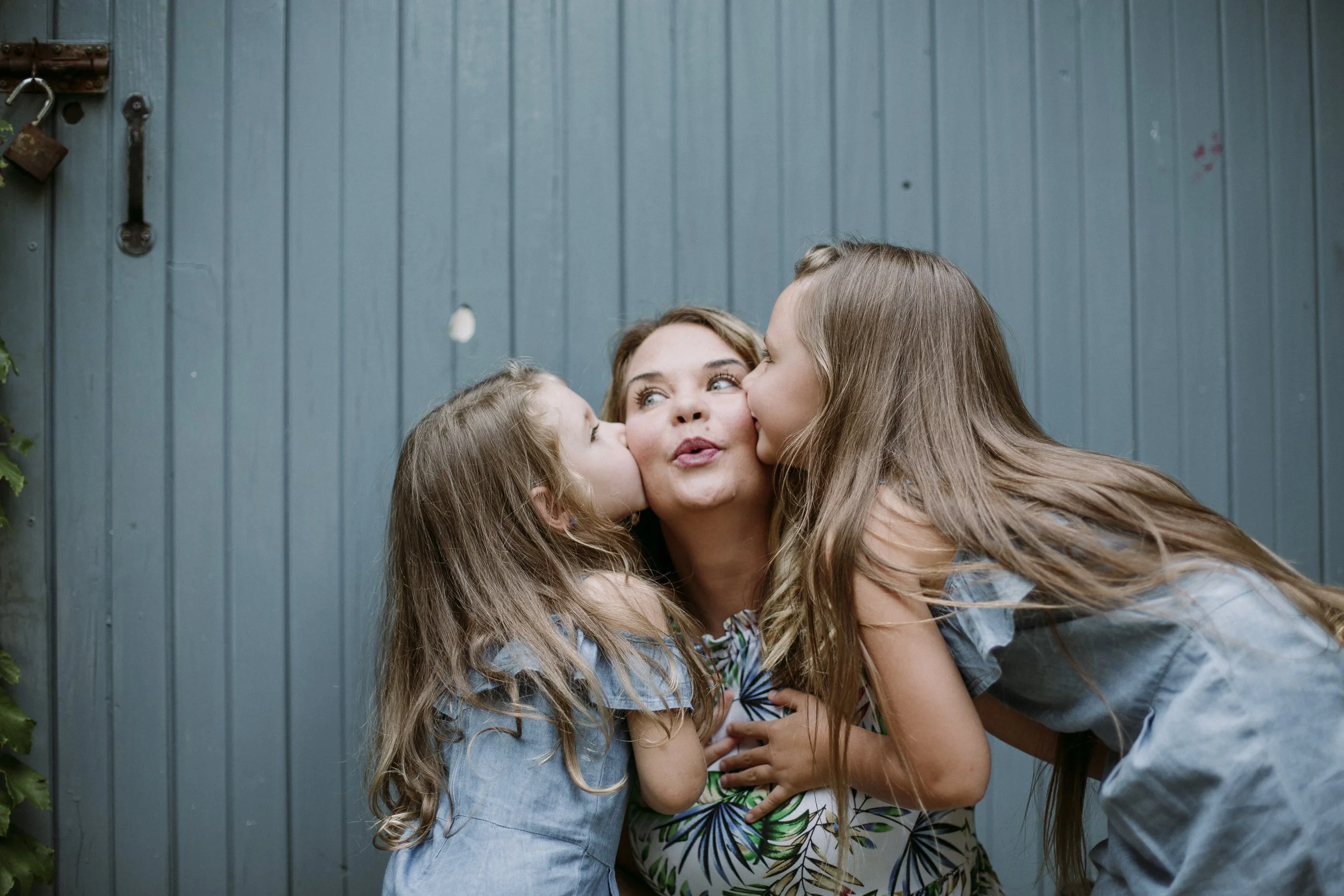 A woman is being kissed on both cheeks by two young girls in front of a blue wooden wall.