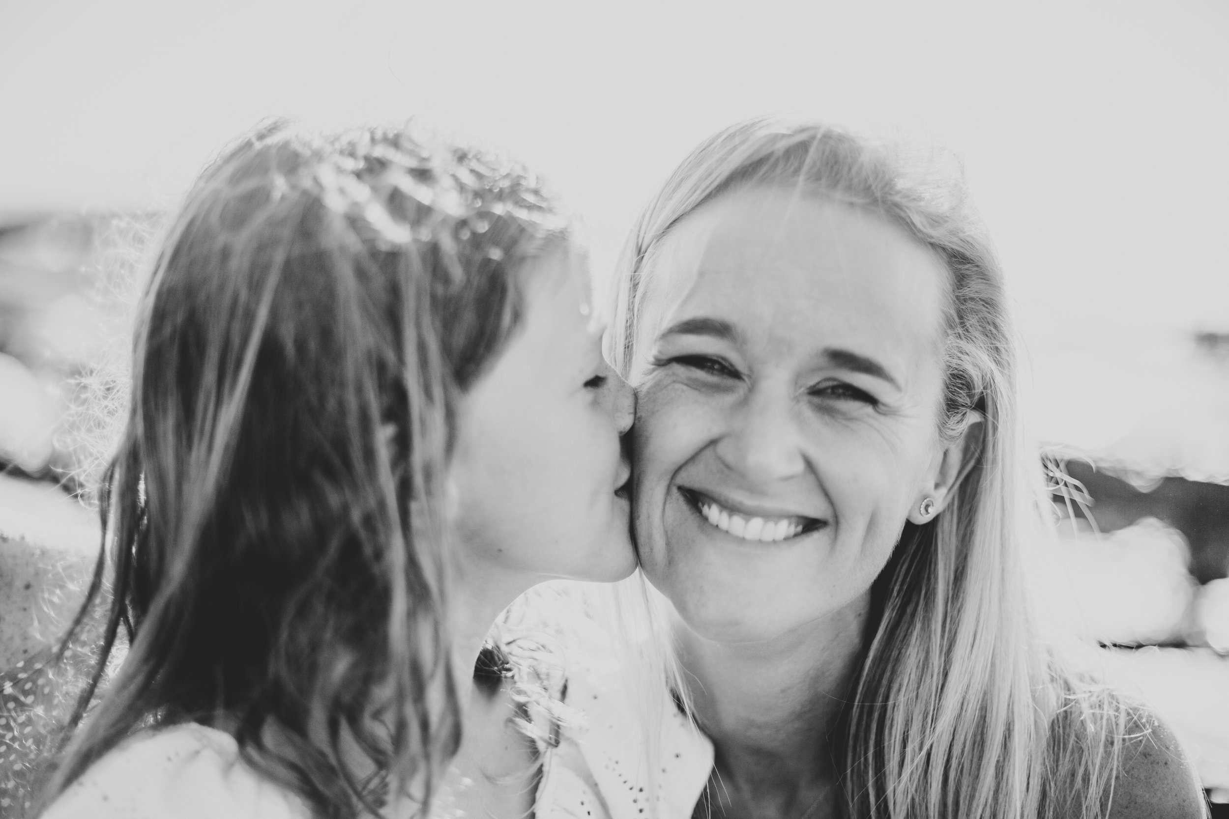 A young girl is giving a woman a kiss on the cheek outdoors, both smiling with the girl’s face turned toward the woman and her eyes closed. Black and white photograph.