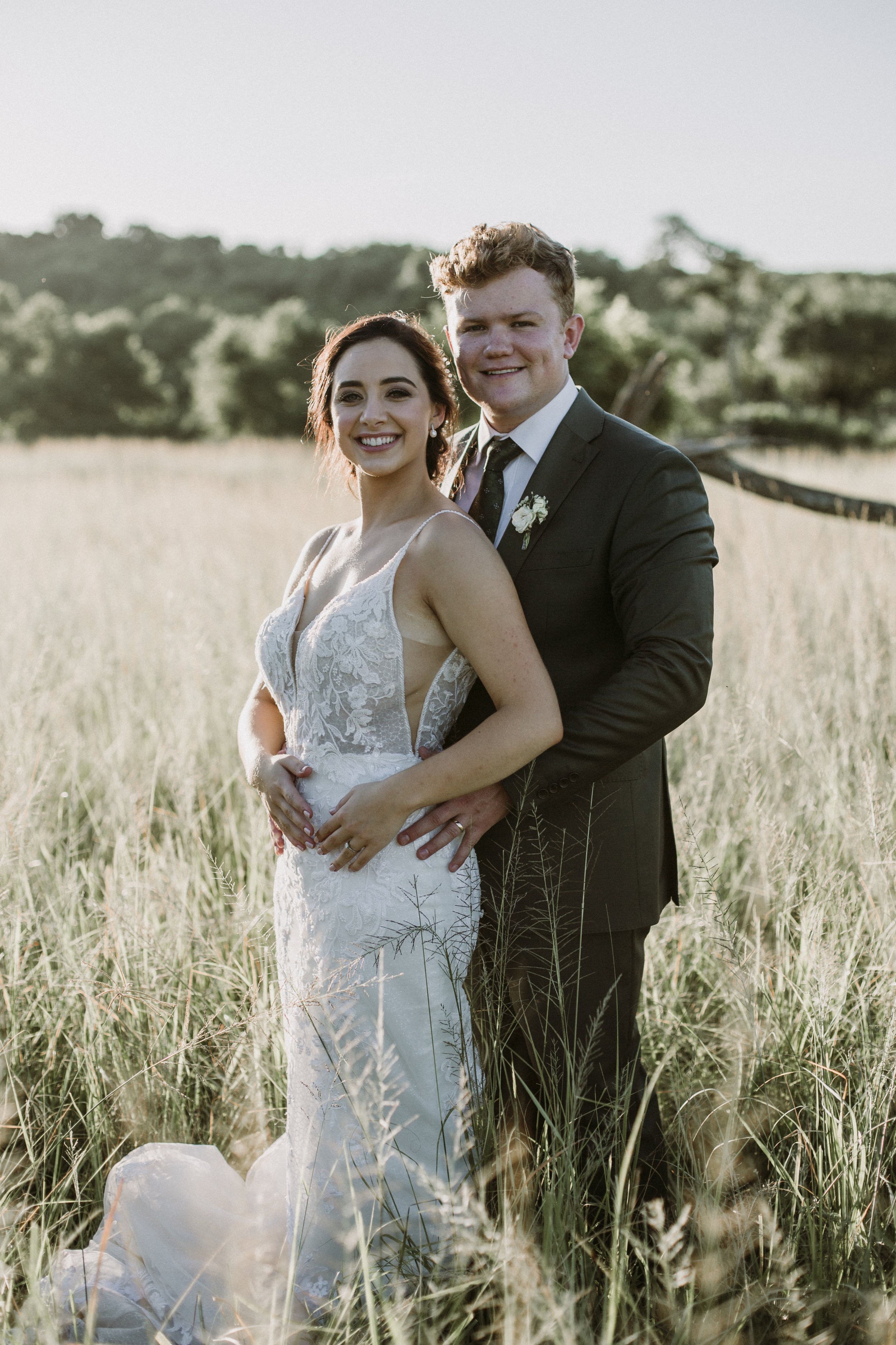 A bride and groom stand together in a field of tall grass, smiling at the camera during their wedding. The bride wears a white lace wedding dress, and the groom is in a dark suit with a tie and boutonniere.