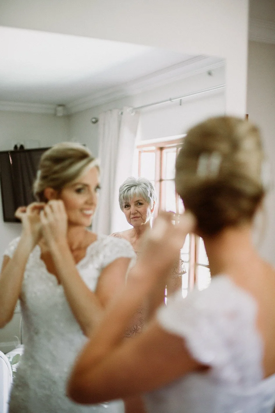 A bride with blonde hair in a white lace dress is putting on earrings while looking at her reflection in a mirror, with an older woman with grey hair watching.