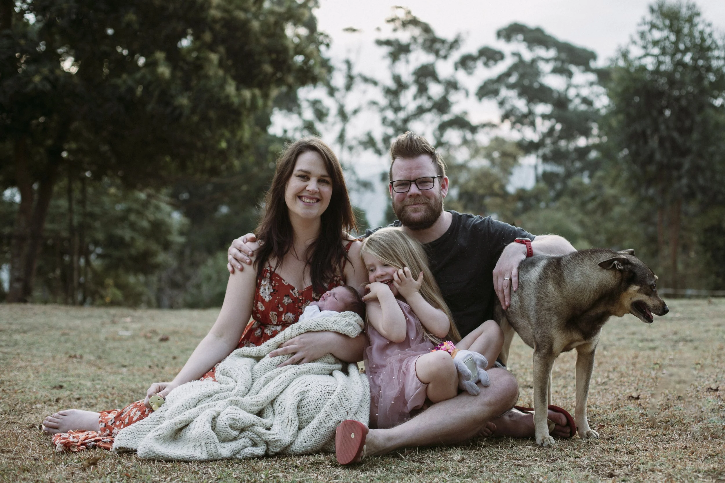 A happy family sitting on the grass outdoors, with two children, a woman, a man, and a dog, in a park with trees in the background.