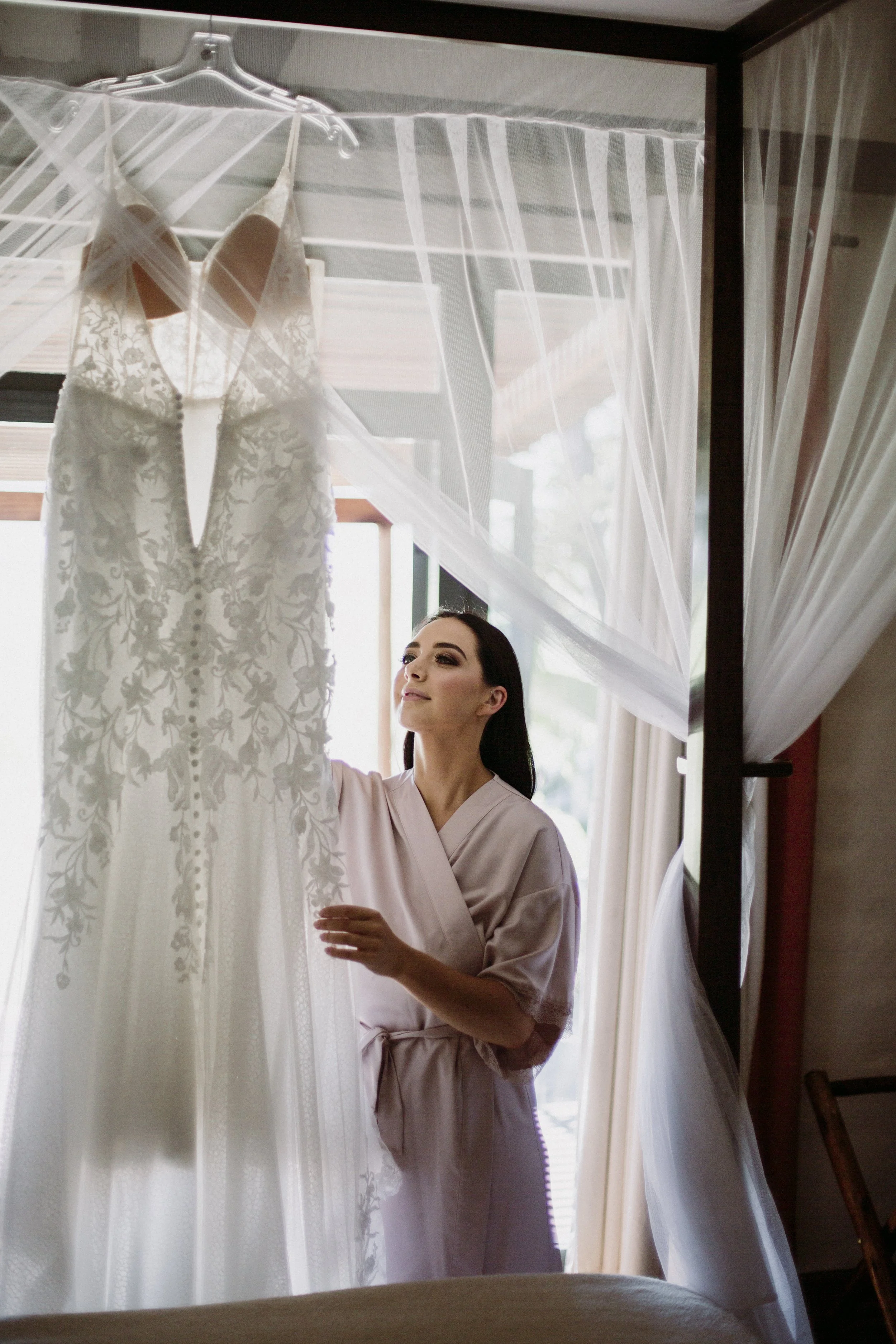 Brunette woman in a pink robe looking at a white lace wedding dress hanging on a canopy bed with sheer curtains.