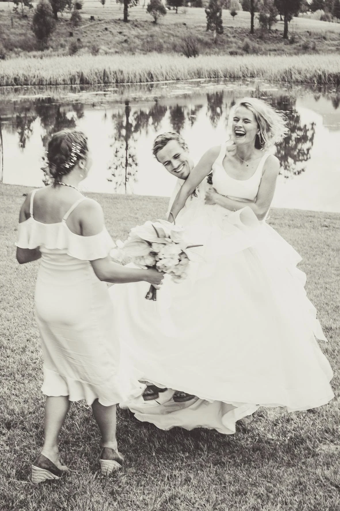 A woman in a wedding dress is laughing as she receives a bouquet of flowers from a girl in a white dress, while a man stands behind her smiling, near a lake with trees in the background.