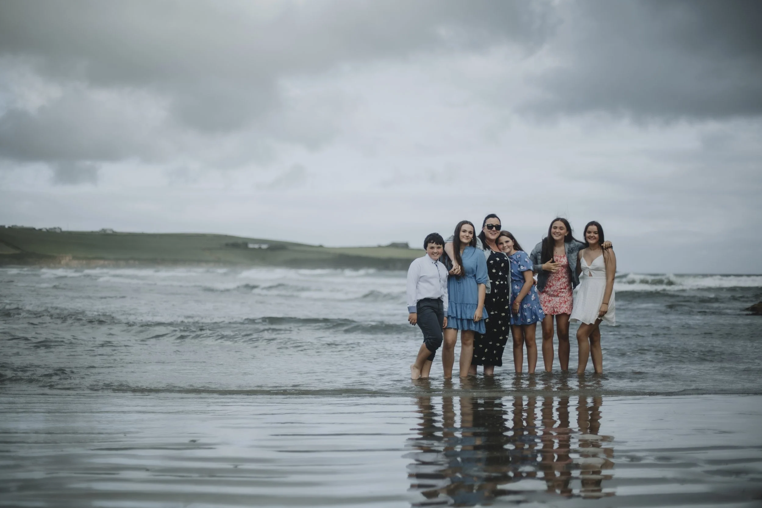 Group of six people standing in shallow ocean water on cloudy day, smiling and posing for a photo.