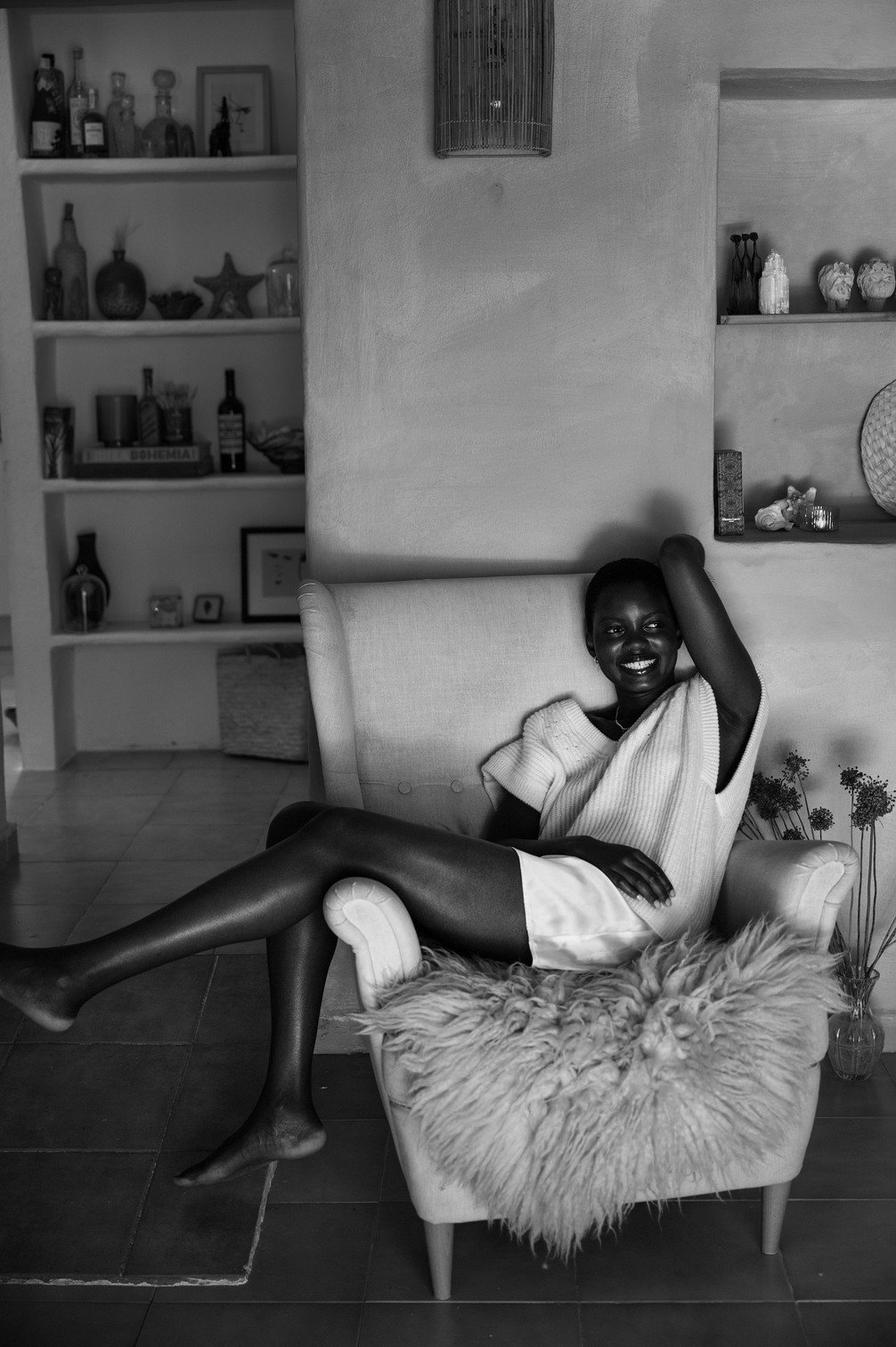 Zwarte vrouw zittend op een gezellige stoel met een pluizige deken, lachend en ontspannen in een huis met decoratieve items en boeken op de achtergrond gefotografeerd door Eric van den Elsen.