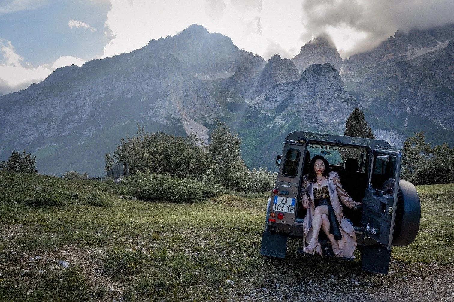 Nazmiye Oral zit op de achterklep van een terreinwagen in een bergachtig landschap met groen gras, struiken en de Alpen in de achtergrond. Ze draagt een jas en een jurk, heeft donker haar en rode lipstick gefotografeerd door Eric van den Elsen.