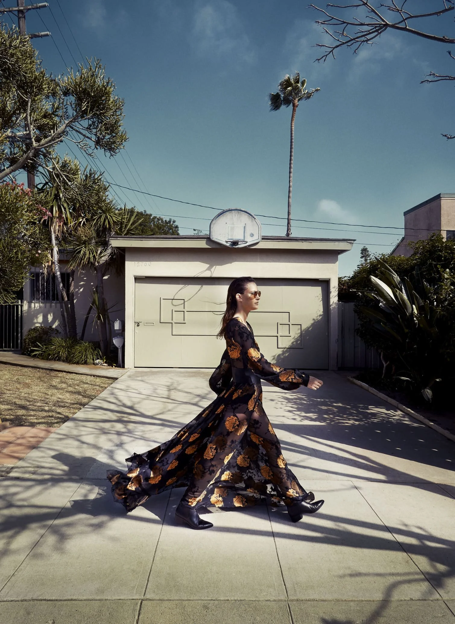 Vrouw in een lange, doorzichtige jurk met bloemenmotief en zittende laarzen wandelt over een parkeerplaats met een garage en een basketballnet op de achtergrond, onder een heldere blauwe hemel met een palmboom gefotografeerd door Eric van den Elsen.