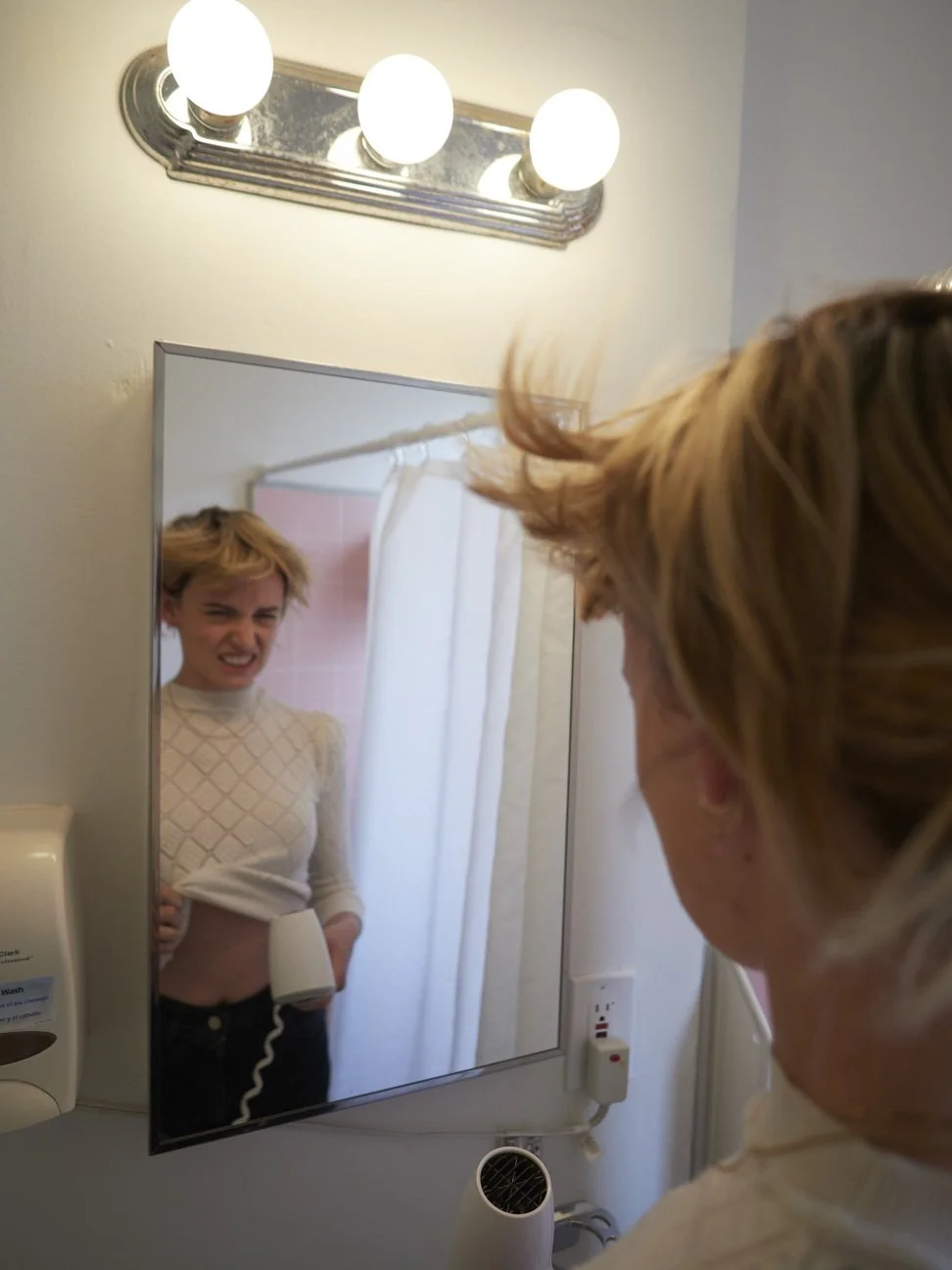Vrouw kijkt in spiegel, die haar uitrekt en frons, terwijl ze haar haar kamt met een föhn in de badkamer gefotografeerd door Eric van den Elsen.