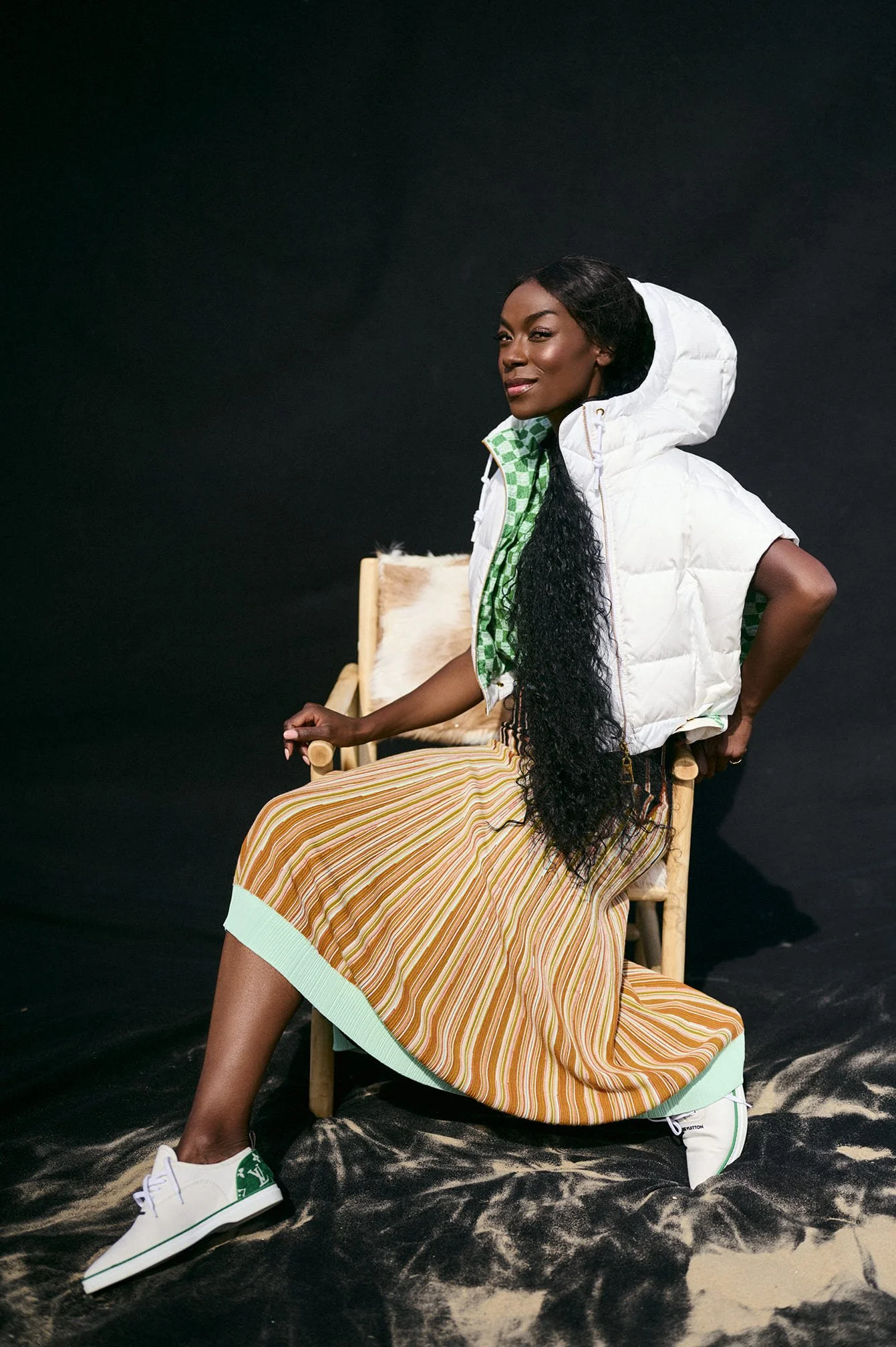 Giovanca Ostiana zittend op een houten stoel tegen een zwarte achtergrond, gekleed in een groene en witte blouse, een lage rok met strepen in aardetinten, een gewatteerde capuchonjas gefotografeerd door Eric van den Elsen.