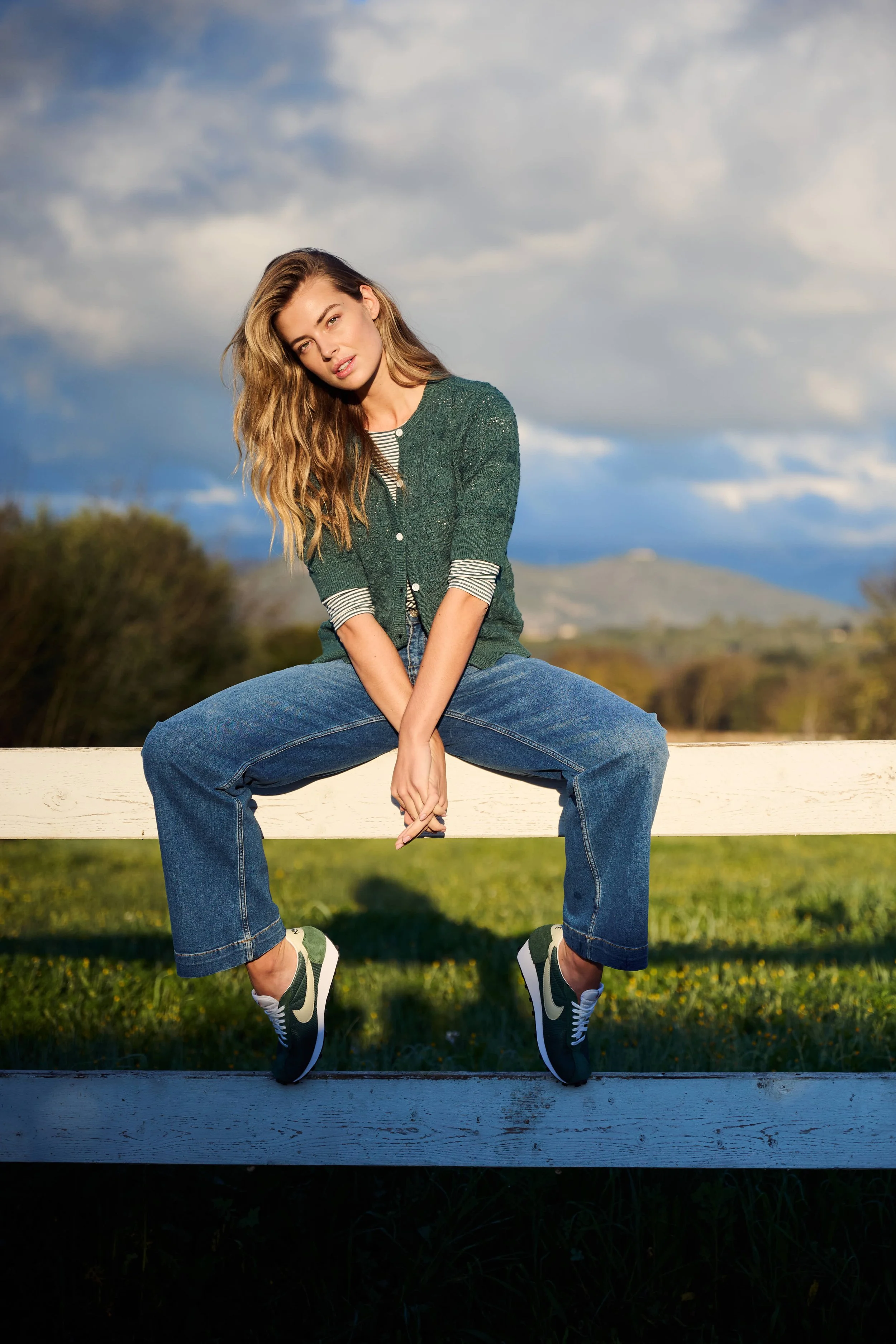 Een jonge vrouw met lang, golvend haar zit op een witte houten hek in een groen veld, gekleed in een groene opengewerkte trui, een gestreepte top, losse blauwe spijkerbroek en sneakers gefotografeerd door Eric van den Elsen voor Josephine & Co.