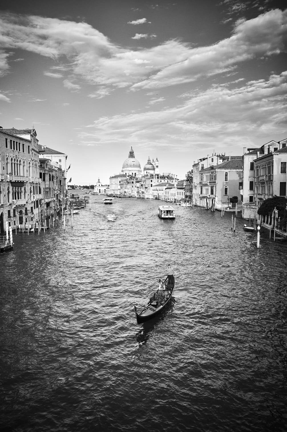 Een zwart-wit foto van een kanaal in Venetië met boten en historische gebouwen aan de oevers, en een grote koepel in de achtergrond onder een bewolkte hemel gefotografeerd door Eric van den Elsen.