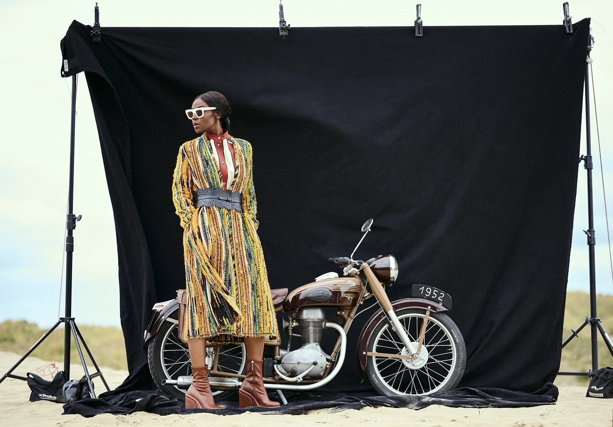 Giovanca Ostiana poseert naast een vintage motorfiets voor een zwarte achtergrond, buiten op het zand, met een blauwachtige lucht en landschap in de verte gefotografeerd door Eric van den Elsen.