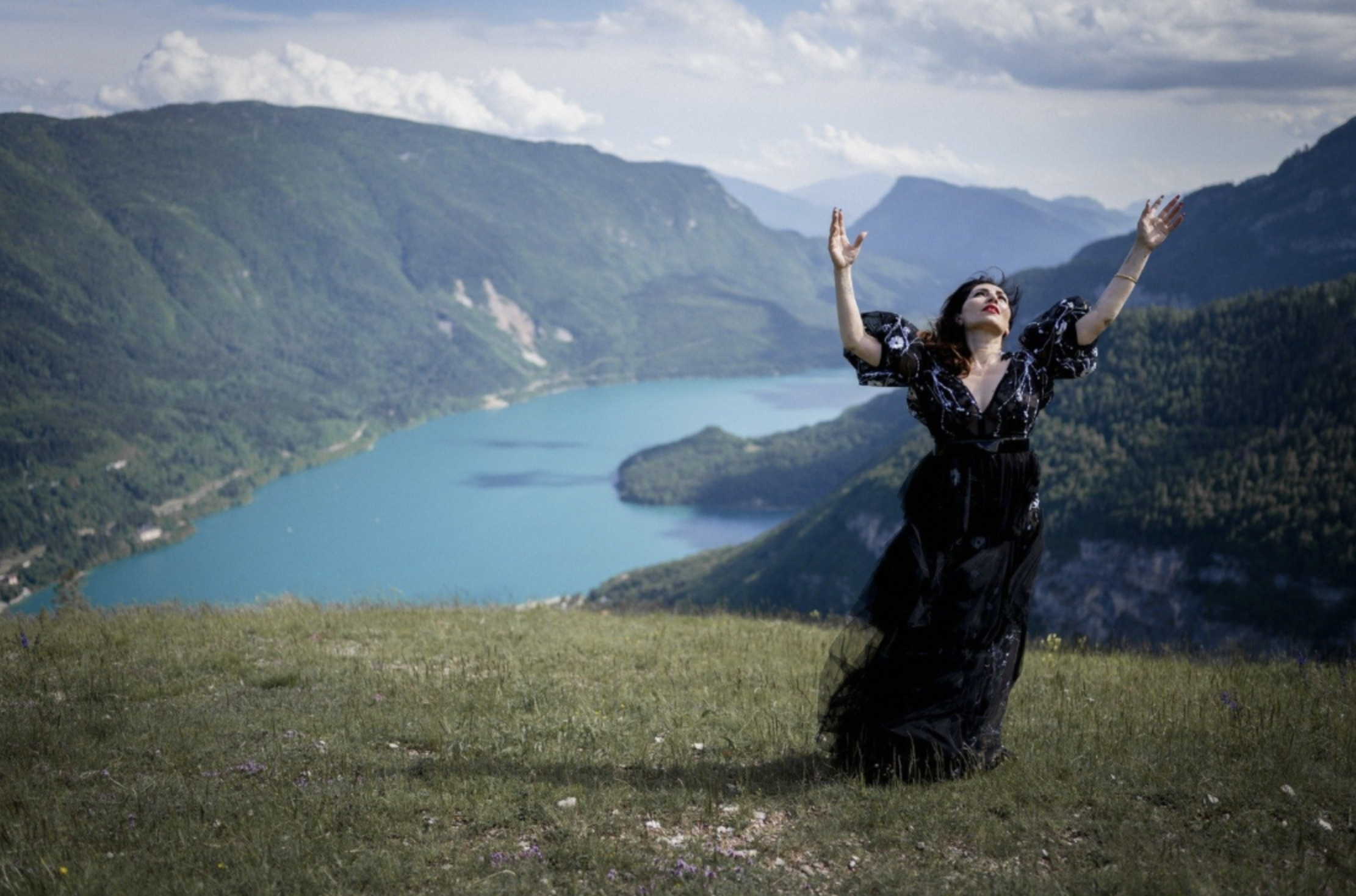 Nazmiye Oral in een zwarte jurk staat met opgetrokken handen op een grasland, met bergen en een meer op de achtergrond gefotografeerd door Eric van den Elsen.
