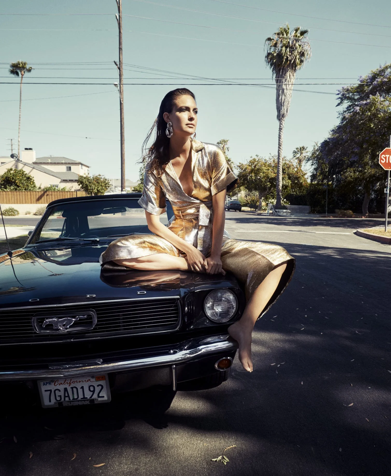 Vrouw in een gouden jurk zit op de motorkap van een zwarte Ford Mustang, met een zonnige achtergrond van palmbomen en een straat gefotografeerd door Eric van den Elsen.