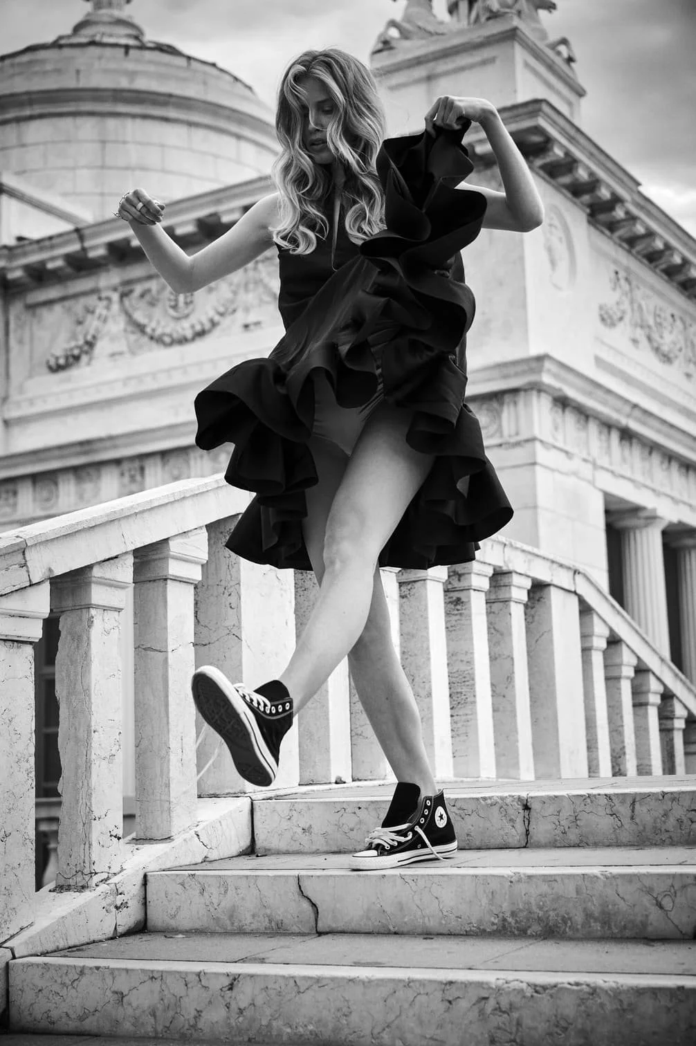 Een vrouw met lang, golvend haar in een zwarte jurk en sneakers staat op een stenen trap voor een oud gebouw met classicale portaattstructuren gefotografeerd door Eric van den Elsen.