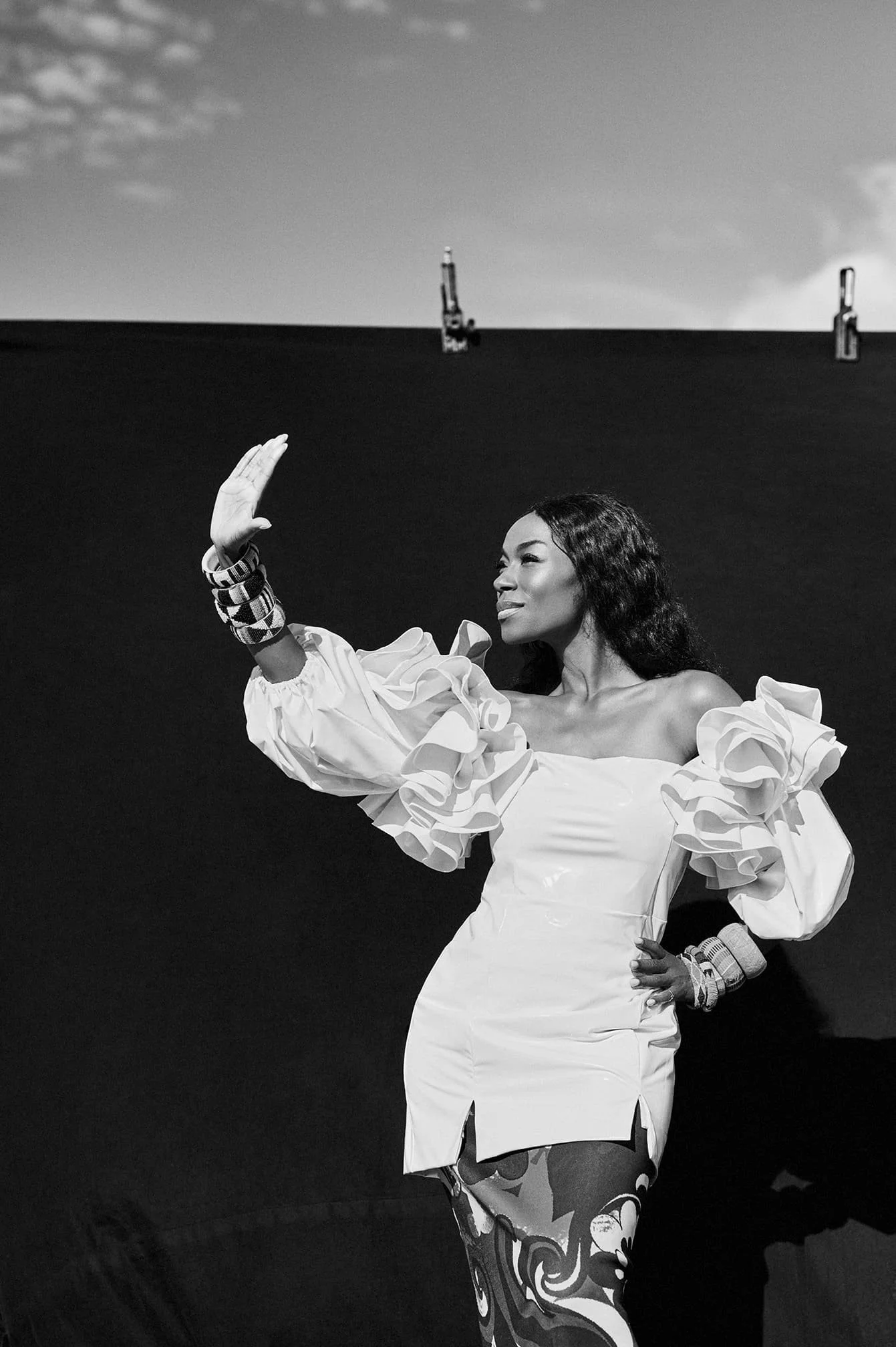 Zwarte en witte foto van Giovanca Ostiana die poseert met een hand omhoog, een grote witte jurk met puffy mouw en opvallende polsaccessoires, achter haar een muur en een schemerige lucht gefotografeerd door Eric van den Elsen.
