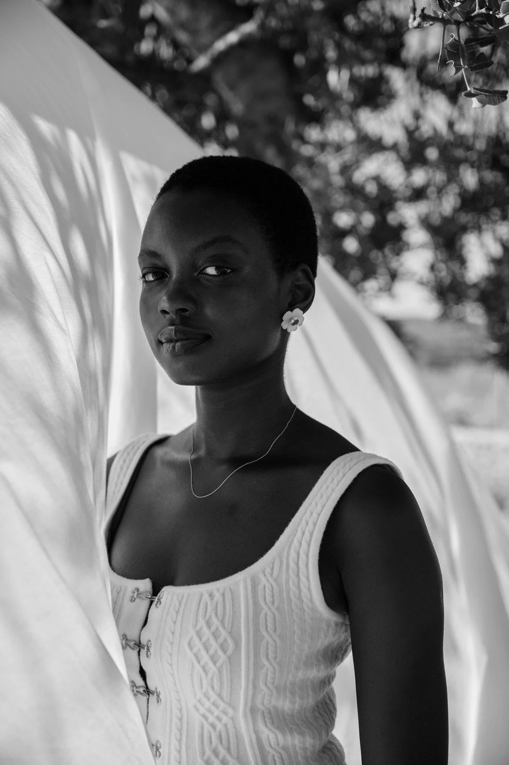 Zwart-wit foto van een vrouw met een korte haardracht, transparant top en oorringen, staand voor een lichte achtergrond met takken en bladeren gefotografeerd door Eric van den Elsen.