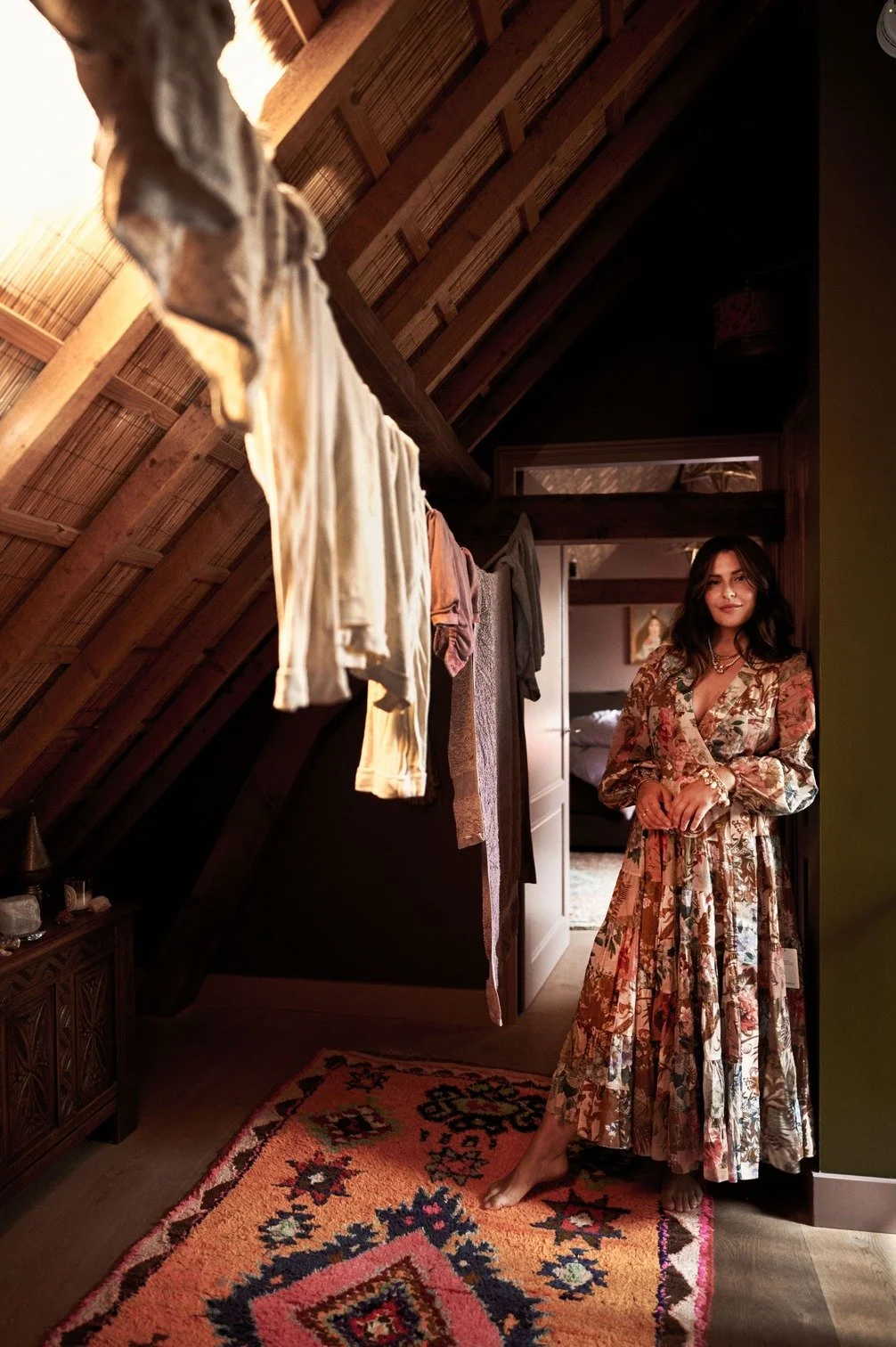 Romy Boomsma in een bloemenjurk staat in een gezellig slaapkamer met een tapijt en kleding die aan een draad hangen, binnen een houten kamer gefotografeerd door Eric van den Elsen.