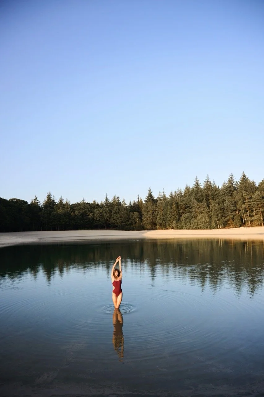 Vrouw in een rode badpak staat in een meer met haar armen omhoog, omringd door een bos en een heldere blauwe hemel gefotografeerd door Eric van den Elsen.