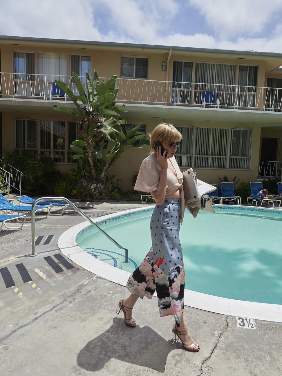 Vrouw met zonnebril, hakken, een vaste coupe en lichte kleding met een lange jurk met een bloemendessin, loopt langs het zwembad terwijl ze belt en documenten vasthoudt gefotografeerd door Eric van den Elsen.